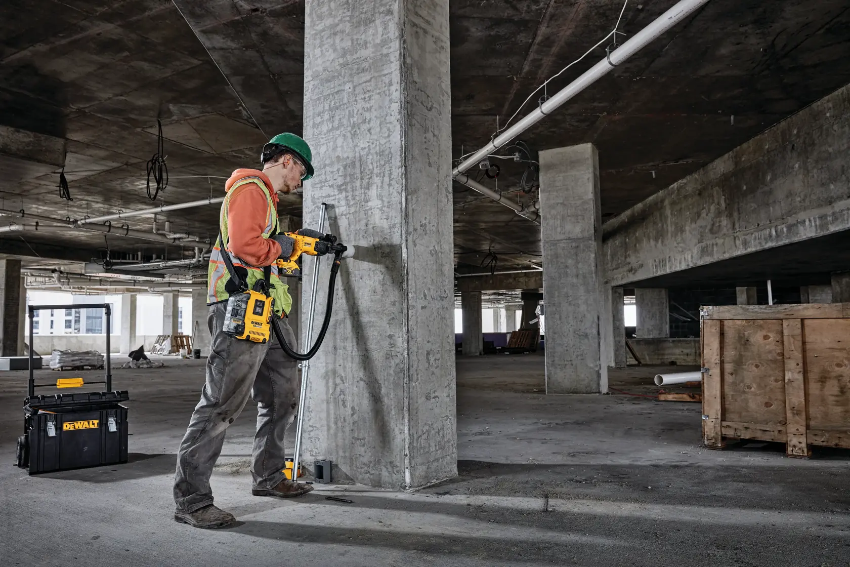 Um trabalhador da construção civil utiliza uma furadeira rotativa DEWALT para perfurar um pilar de concreto dentro de um edifício parcialmente construído. Próximo, há uma caixa de ferramentas DEWALT no chão.