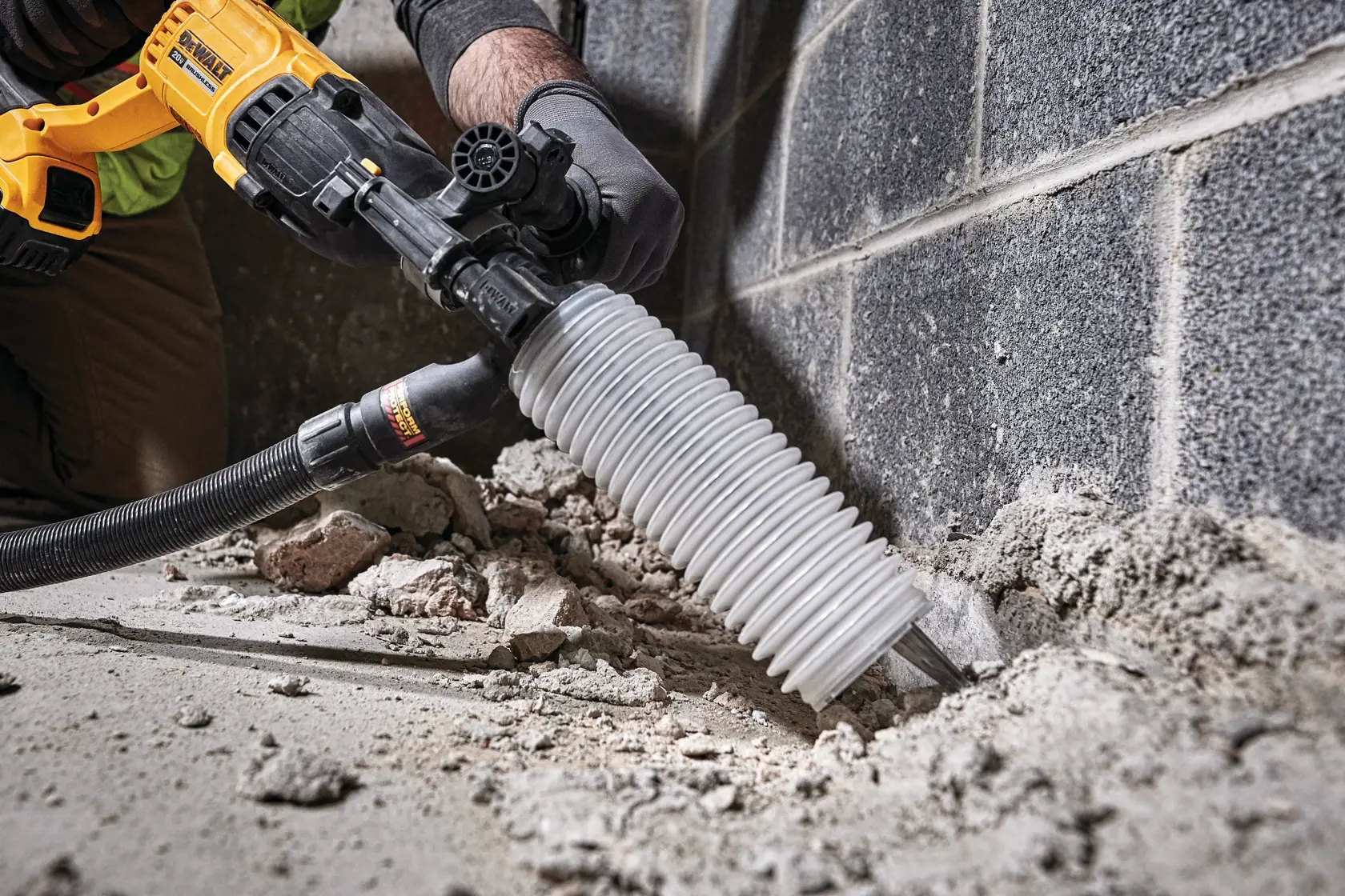 Uma pessoa está usando um martelo rotativo DEWALT com um acessório de coleta de poeira para quebrar concreto próximo a uma parede. A ferramenta é amarela e preta, e há destroços visíveis no chão.