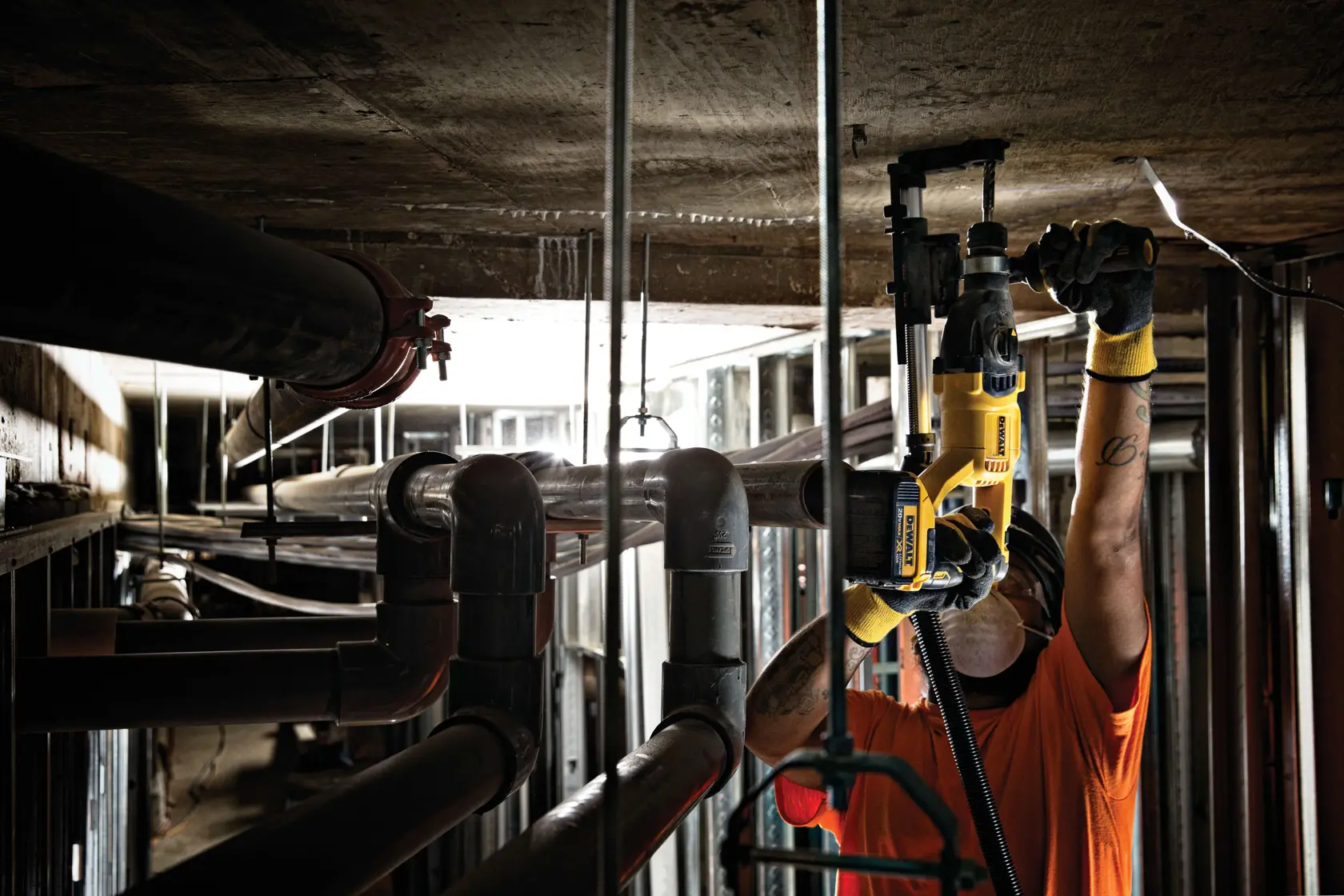 Uma pessoa usando capacete de segurança e luvas utiliza um martelete rotativo amarelo DEWALT para trabalhar em um teto de concreto em um ambiente industrial com tubos e estruturas metálicas visíveis.
