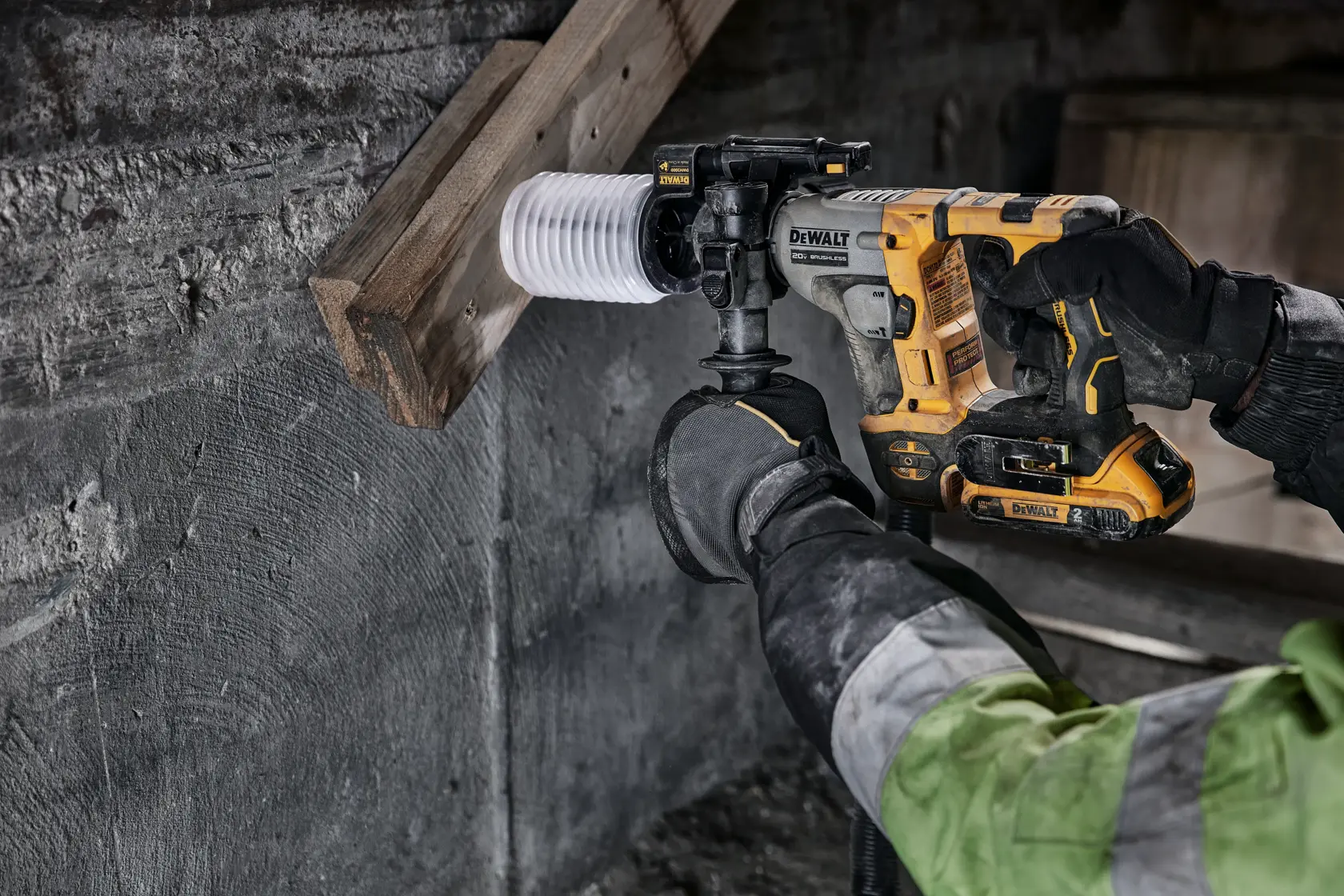 Uma pessoa está usando um martelete rotativo sem fio DEWALT para perfurar um buraco em uma viga de madeira e uma parede de concreto. O usuário está usando luvas e uma jaqueta de trabalho.