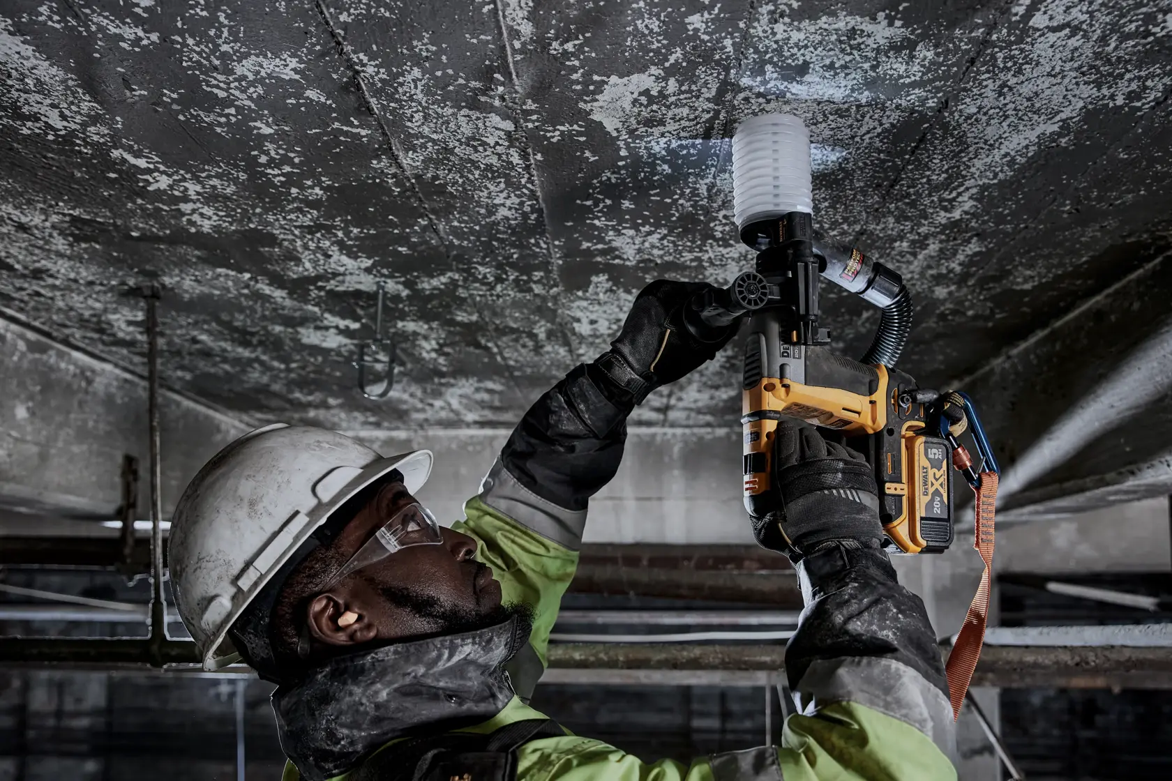 Um trabalhador usando equipamentos de segurança, incluindo capacete, luvas e óculos de proteção, utiliza uma ferramenta elétrica amarela DEWALT para perfurar um teto de concreto em um ambiente industrial.