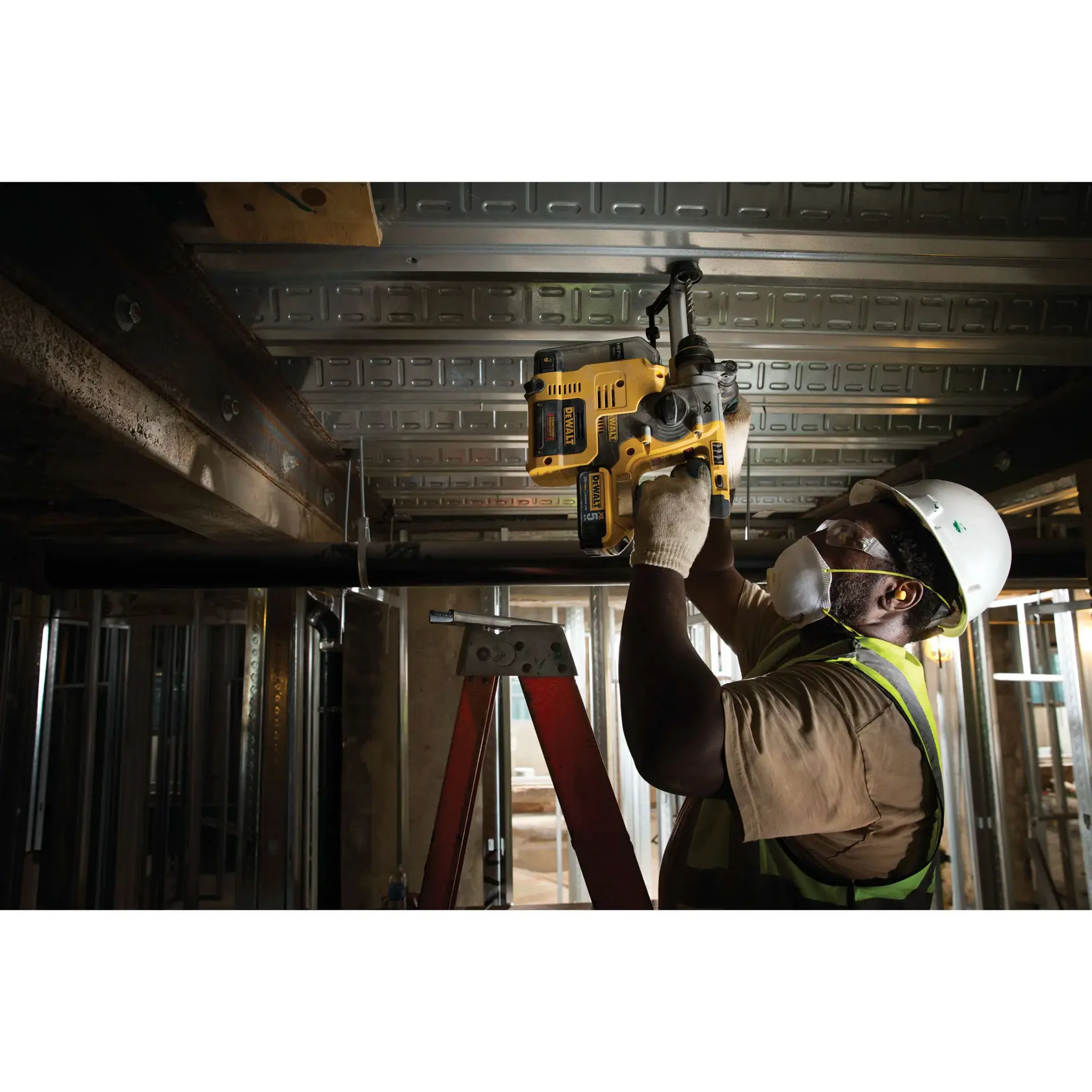 Um trabalhador da construção civil usando capacete, colete de segurança, luvas e máscara está utilizando uma furadeira rotativa amarela DEWALT para trabalhar no teto de metal, estando em cima de uma escada em um ambiente industrial.