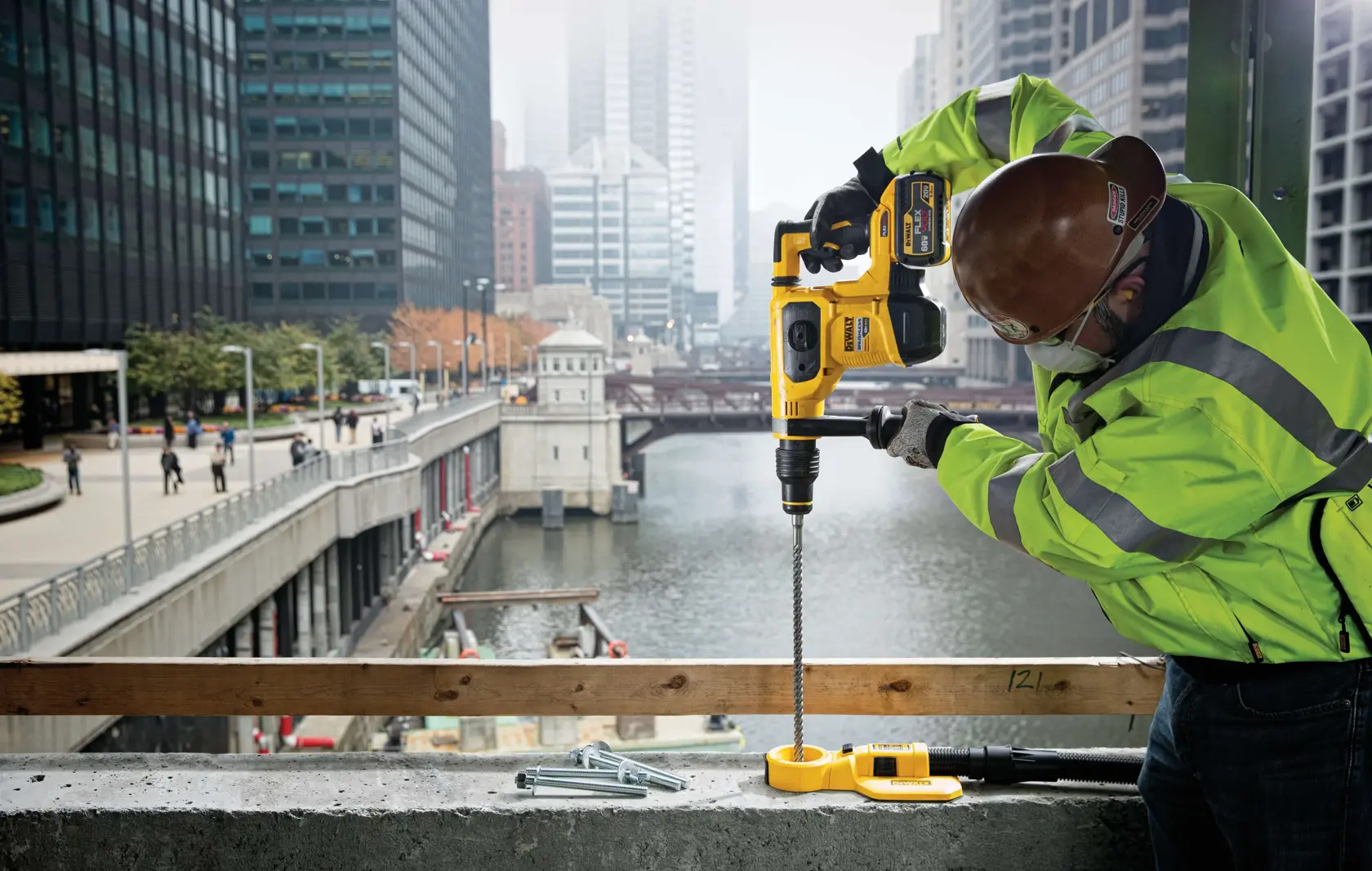 Um trabalhador da construção civil com capacete de segurança e jaqueta de alta visibilidade usa um martelete DEWALT (SKU: DCH481X2) para perfurar concreto em um canteiro de obras próximo a um rio, com arranha-céus ao fundo.