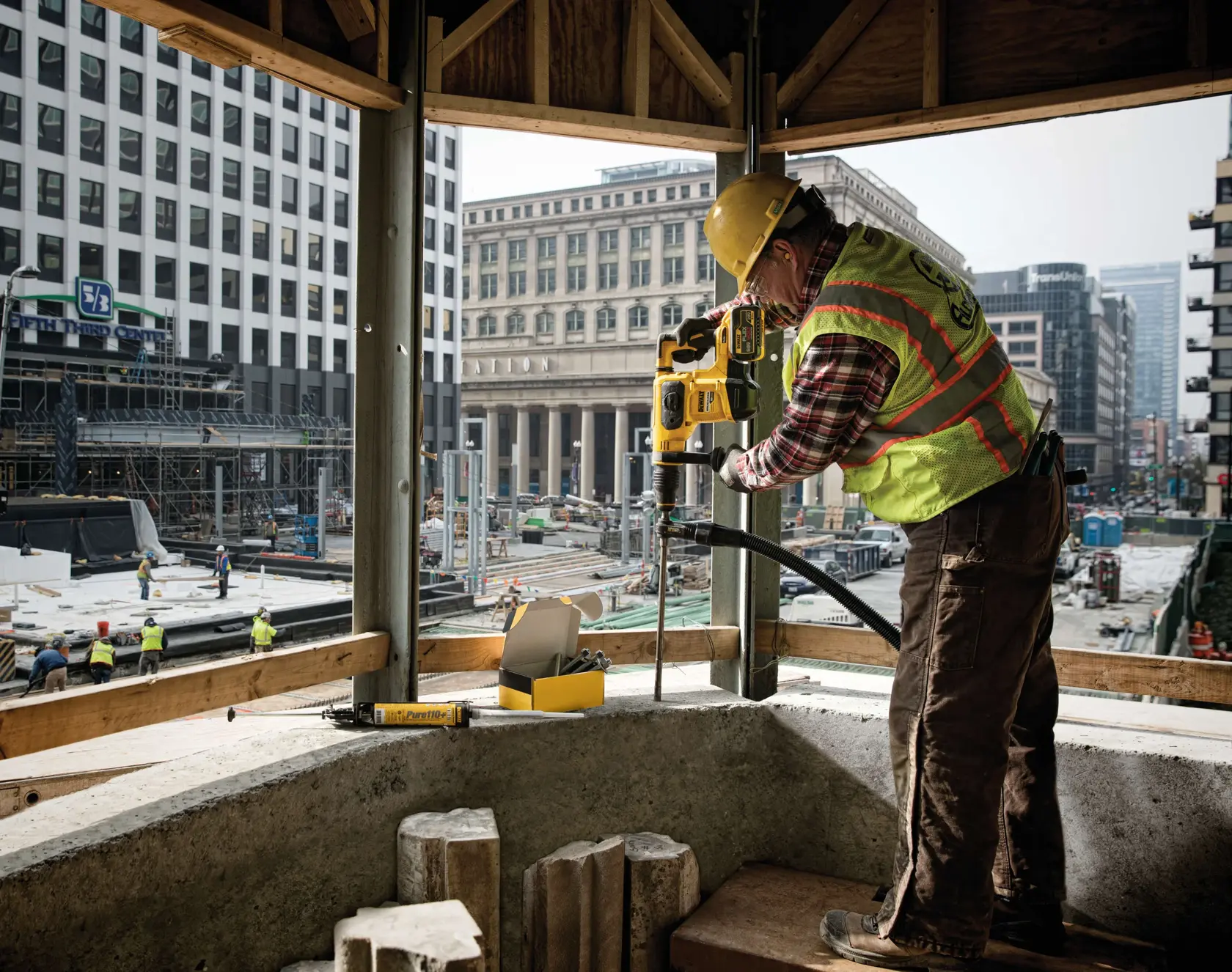 Um trabalhador da construção civil utiliza uma furadeira rotativa DEWALT (DCH481X2) para perfurar uma superfície de concreto em um canteiro de obras. O trabalhador está usando um capacete amarelo e um colete refletivo. A cena é ao ar livre, em uma área urbana com edifícios altos ao fundo. Acessórios e embalagens de ferramentas DEWALT estão próximos na plataforma.