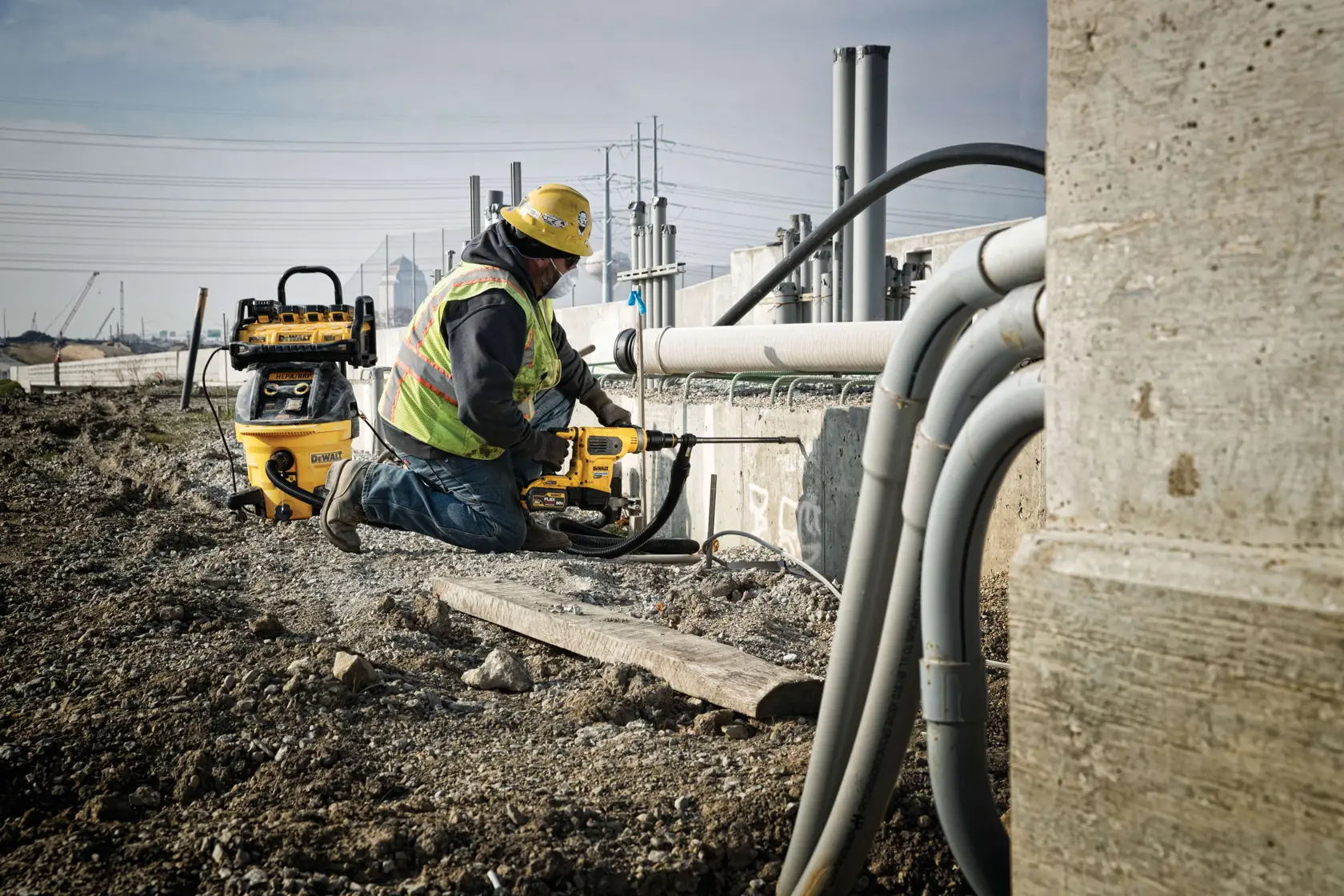 Um trabalhador usando colete de segurança e capacete utiliza uma ferramenta elétrica DEWALT em um canteiro de obras, ajoelhado ao lado de concreto e tubos. Equipamentos adicionais da DEWALT são visíveis ao fundo.