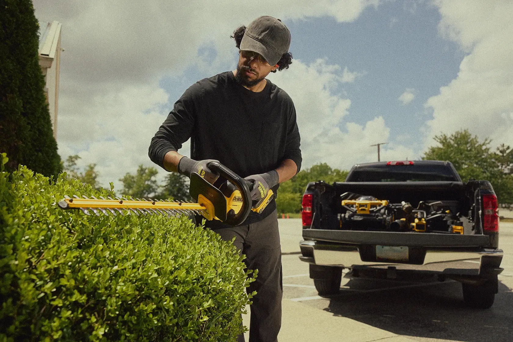 Uma pessoa com o rosto desfocado usa um aparador de cerca viva sem fio DEWALT para podar arbustos ao ar livre. A pessoa está usando luvas e boné. Ao fundo, uma caminhonete com ferramentas DEWALT na caçamba aberta está estacionada.