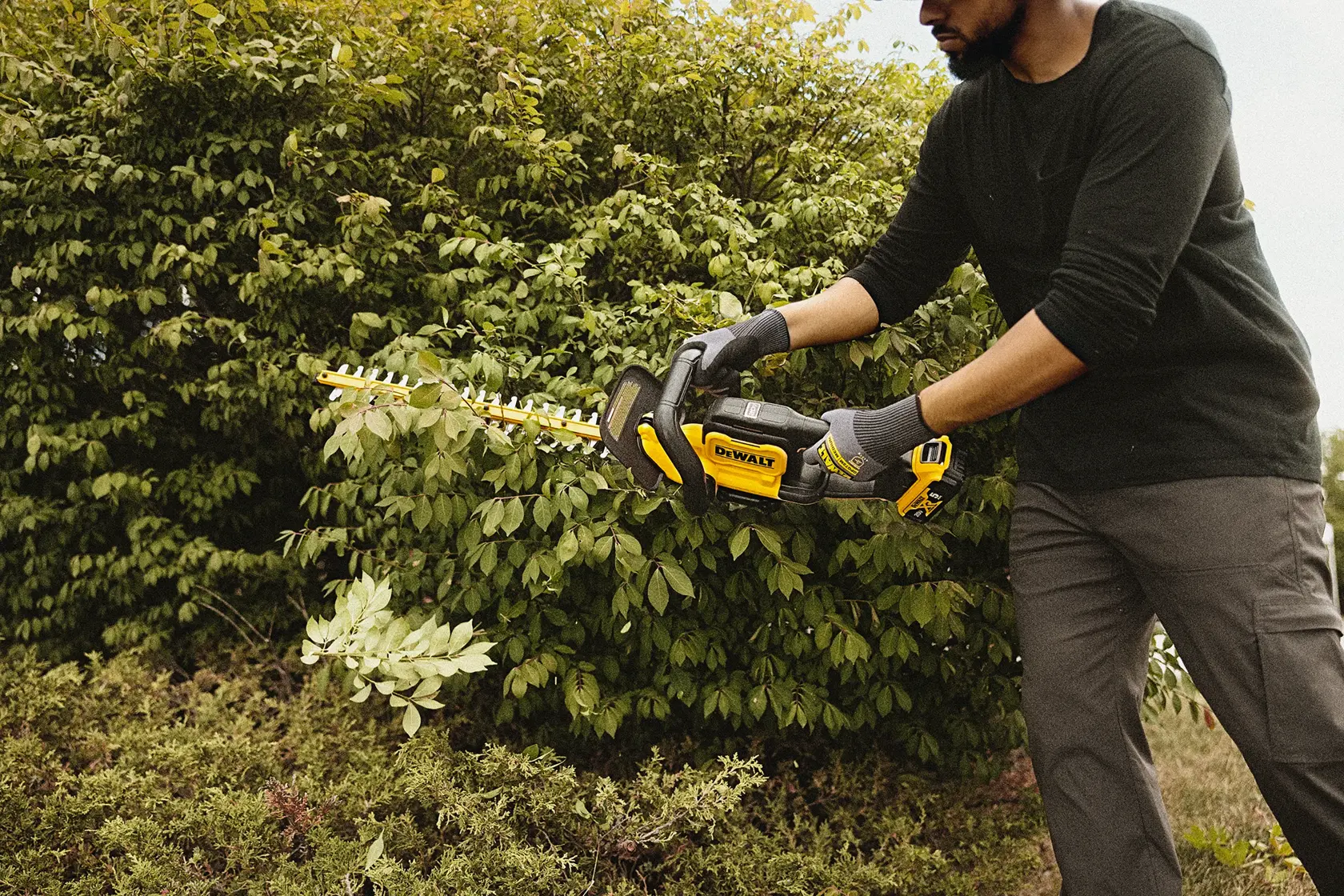 Uma pessoa usando luvas utiliza um aparador de cerca viva sem fio DEWALT DCHT821B para podar arbustos ao ar livre. O rosto da pessoa está desfocado para garantir a privacidade.