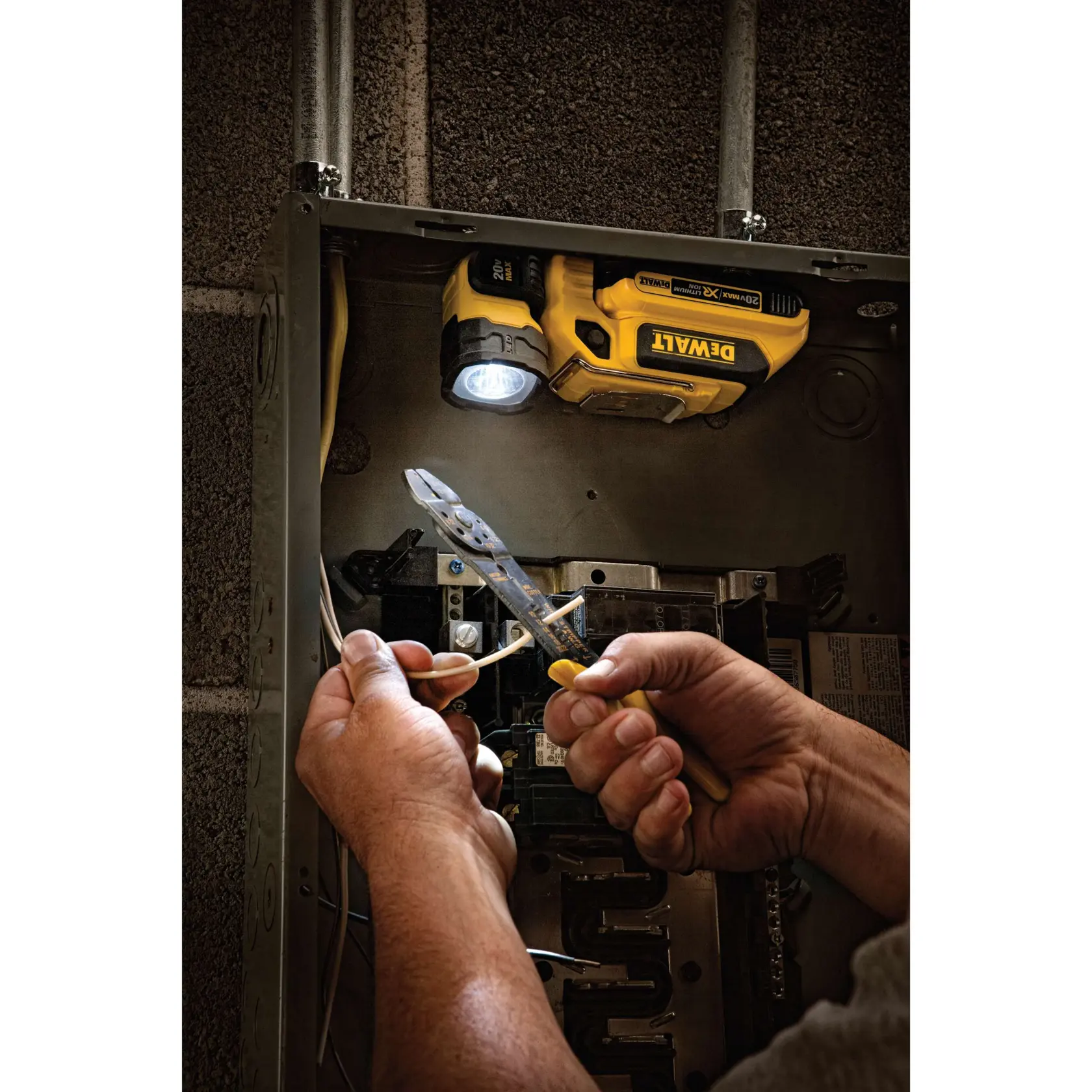 Uma luz de trabalho LED portátil DEWALT (modelo DCL044) ilumina o interior de um painel elétrico enquanto mãos utilizam um alicate para descascar fios.