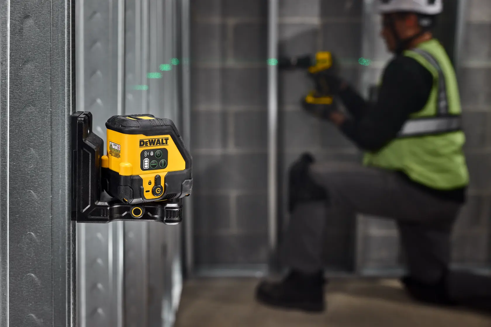 Uma ferramenta de nível a laser DEWALT montada em uma estrutura de parede metálica, projetando uma linha de laser verde pela sala. Ao fundo, uma pessoa usando capacete de segurança e colete refletivo trabalha com outra ferramenta DEWALT, ajoelhada perto da estrutura metálica.
