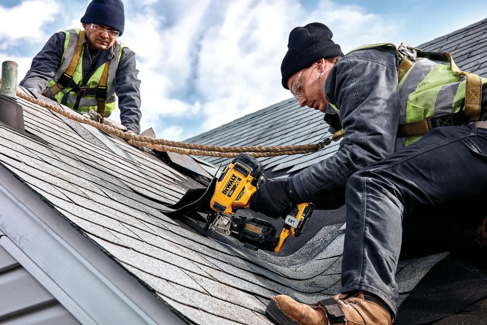 Dois trabalhadores da construção usando equipamentos de segurança e cintos estão trabalhando em um telhado com telhas. Um deles utiliza uma pregadora de telhado sem fio DEWALT DCN45RNB para fixar as telhas, enquanto o outro auxilia. O céu está parcialmente nublado ao fundo.