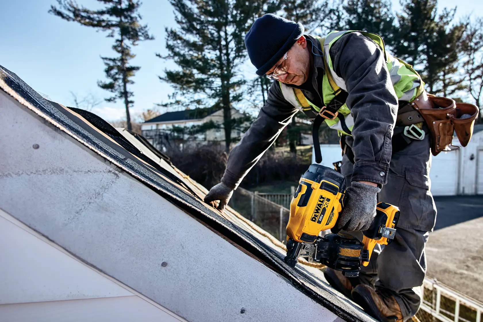 Um trabalhador usando uma pregadora de telhado DEWALT DCN45RNB em um telhado inclinado. O trabalhador está usando equipamento de segurança e luvas, e está fixando telhas no telhado. O fundo inclui árvores e uma área residencial.