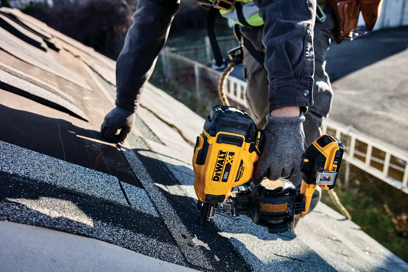 Uma pessoa com luvas de trabalho está usando uma pregadora de telhado sem fio DEWALT em um telhado de telhas. A ferramenta é amarela e preta, e a pessoa está fixando telhas enquanto trabalha em um telhado inclinado.