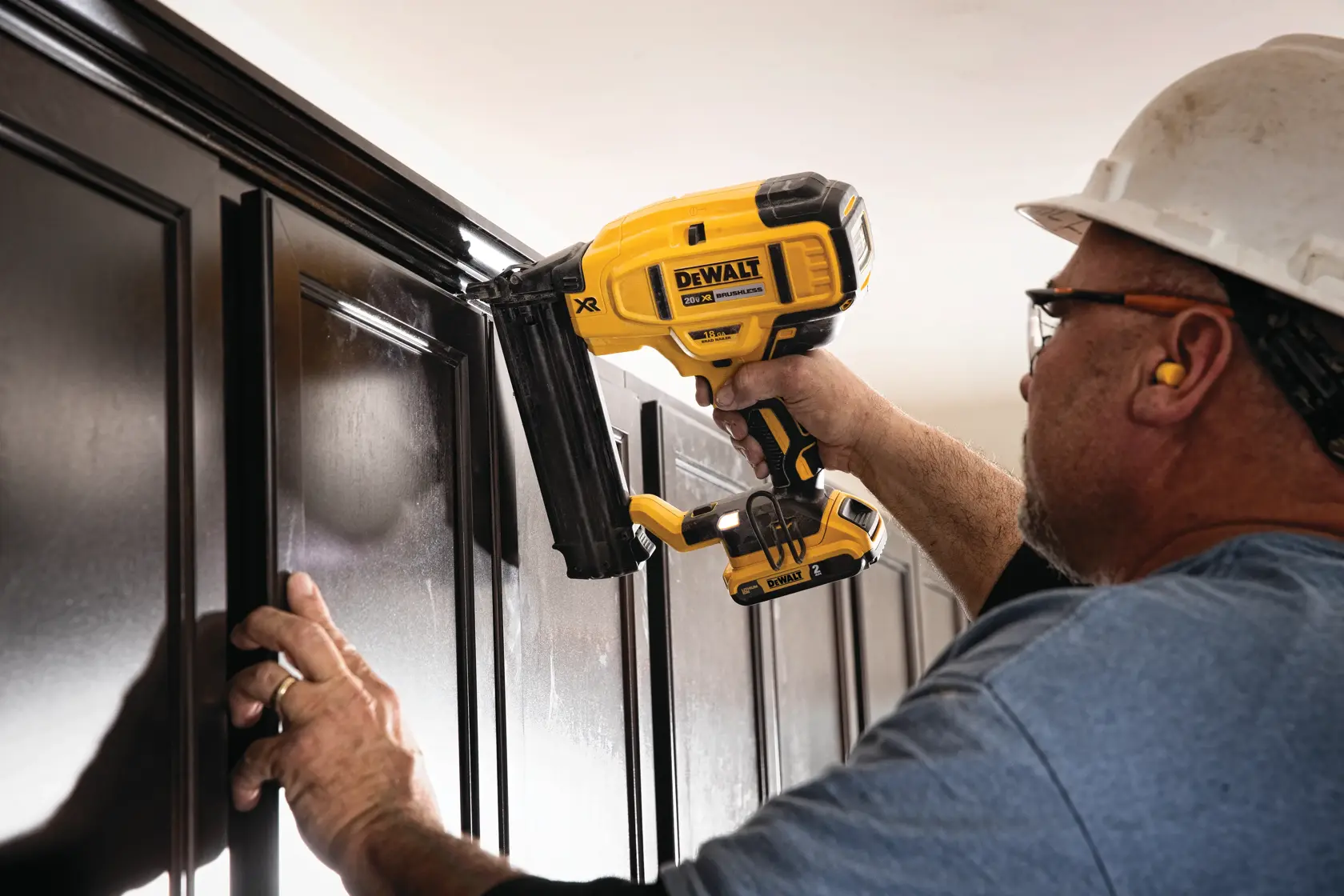 Uma pessoa usando equipamentos de segurança utiliza uma pregadora sem fio DEWALT para instalar acabamento em armários de madeira escura.