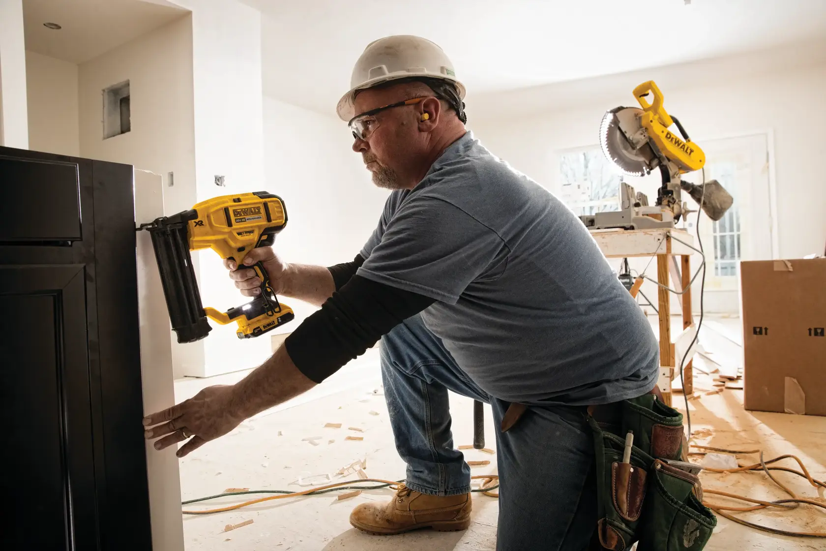Uma pessoa usando capacete e proteção auricular está utilizando uma pistola de pregos amarela DEWALT (modelo DCN680B) para fixar um armário ou painel em ambiente interno. A pessoa está ajoelhada no chão em um local de construção ou reforma, com aparas de madeira e ferramentas elétricas visíveis ao fundo.