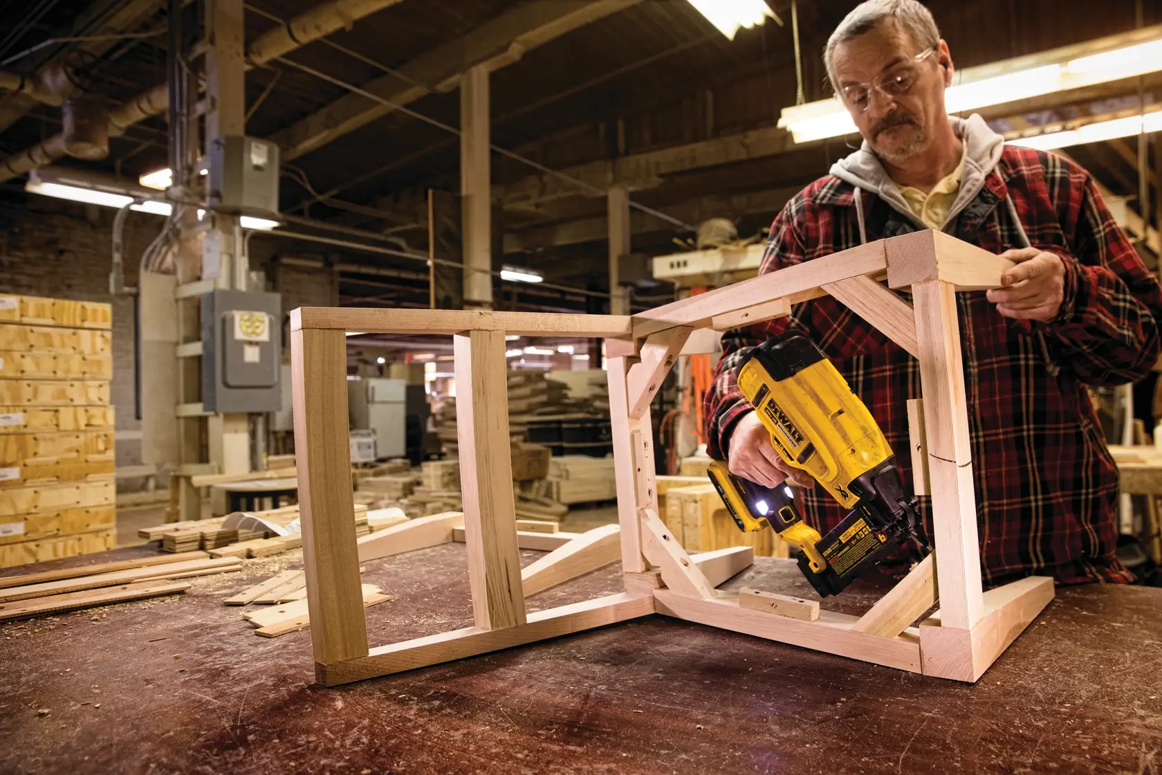 Uma pessoa utiliza uma pregadora DEWALT DCN680B para montar uma estrutura de madeira em uma oficina. O rosto da pessoa está desfocado e ela está vestindo uma camisa xadrez. O espaço de trabalho está cheio de madeira e equipamentos de oficina.