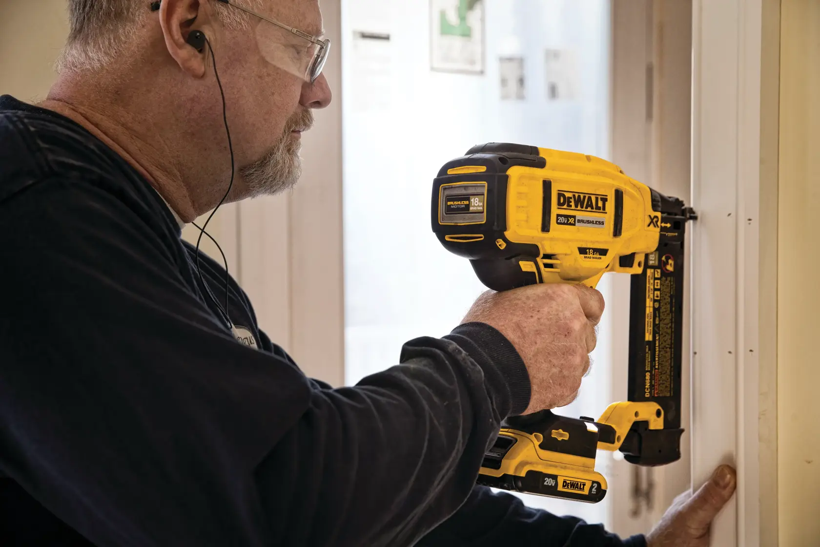 Uma pessoa utiliza uma pregadora sem fio DEWALT DCN680B para fixar uma moldura em uma porta. A ferramenta amarela e preta, de marca registrada, é segurada com uma mão, e o rosto do indivíduo está desfocado para garantir a privacidade.