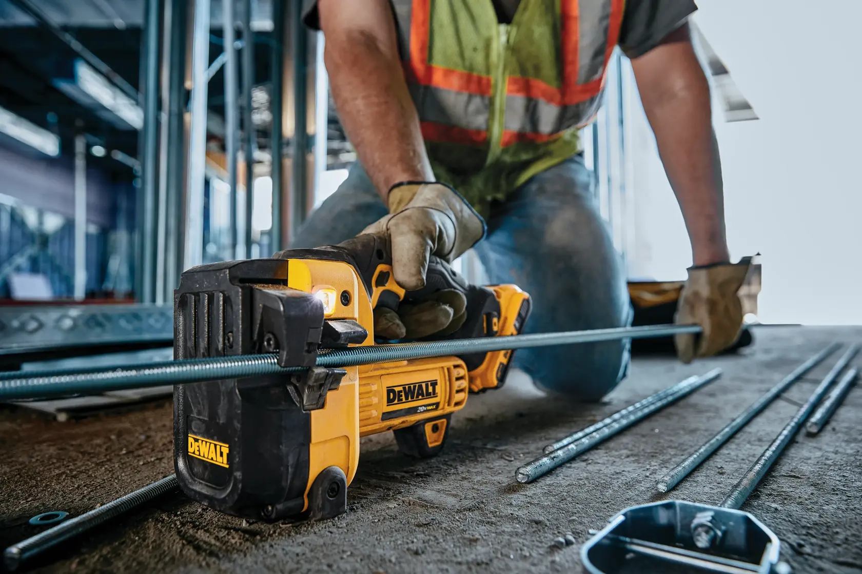 Uma pessoa usando luvas de trabalho e colete de segurança utiliza uma ferramenta de corte sem fio DEWALT para cortar uma barra de metal em um canteiro de obras.