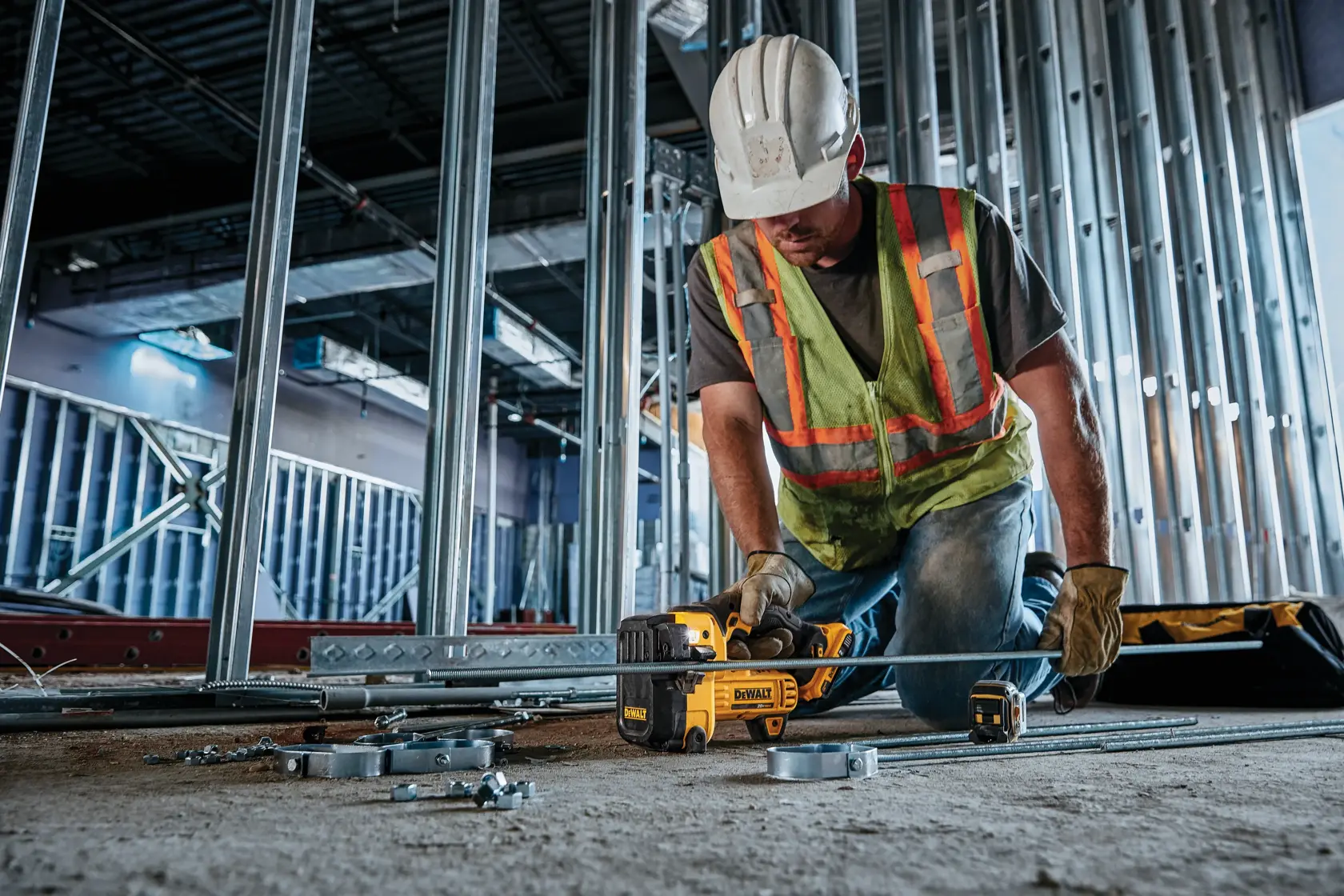 Um trabalhador da construção civil usando colete de segurança, luvas e capacete utiliza uma ferramenta DEWALT DCS350B para cortar barras de metal em um canteiro de obras, cercado por estruturas metálicas e suprimentos de construção.