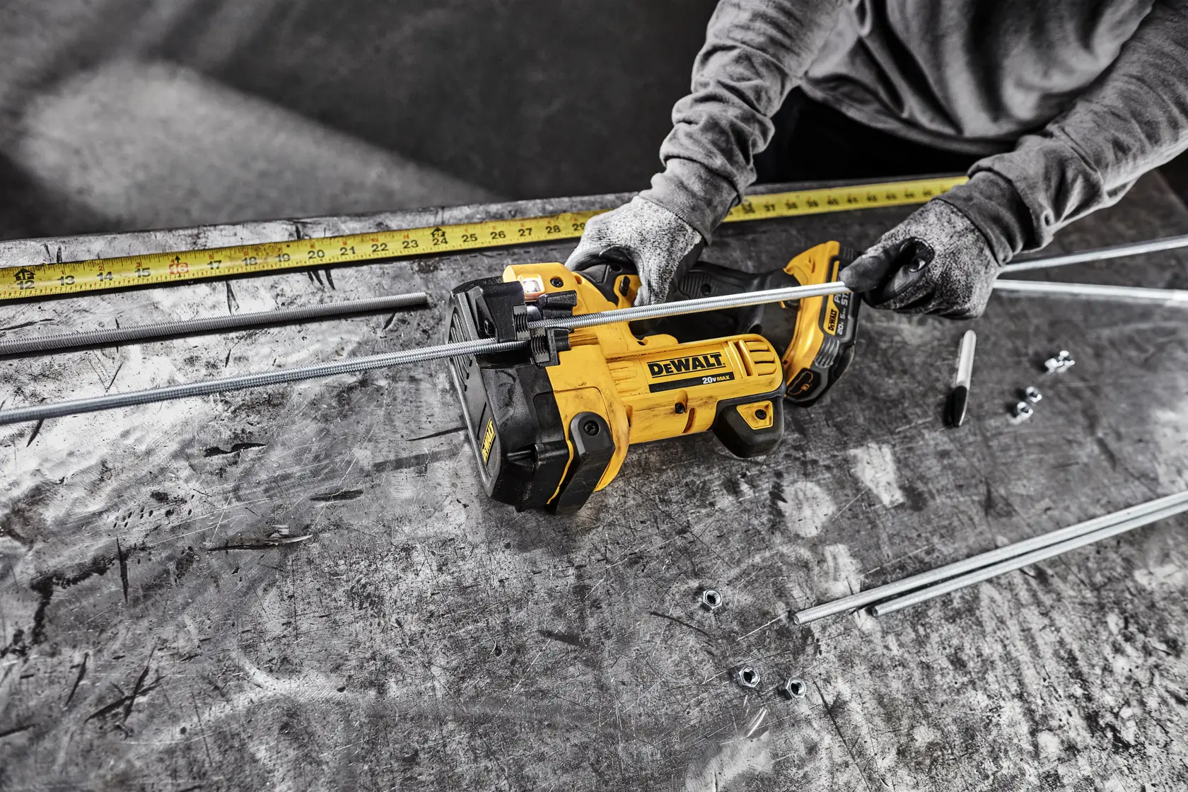 Uma pessoa usando luvas utiliza um cortador de haste roscada sem fio amarelo DEWALT para cortar hastes metálicas em uma bancada de trabalho. Várias hastes, porcas e uma fita métrica estão visíveis na mesa.