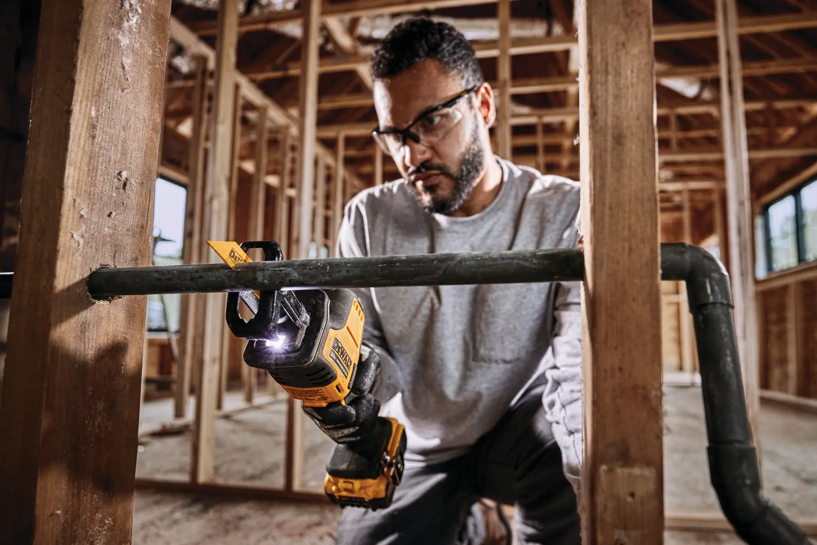 Uma pessoa usando uma serra recíproca sem fio DEWALT para cortar um tubo de metal em um ambiente de construção, com luvas de segurança e estrutura de madeira visíveis ao fundo. O rosto da pessoa está borrado.