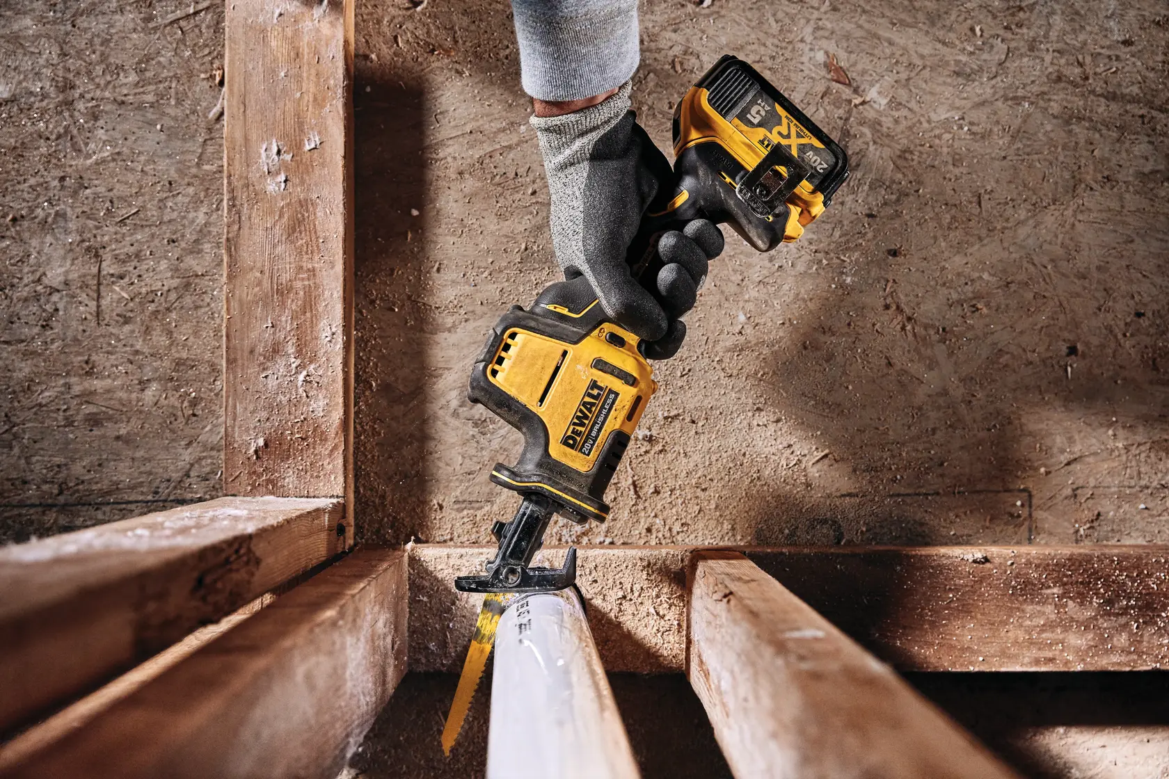 Uma mão com luva está usando uma serra recíproca sem fio amarela e preta da DEWALT para cortar um tubo de plástico branco em uma estrutura de construção de madeira.