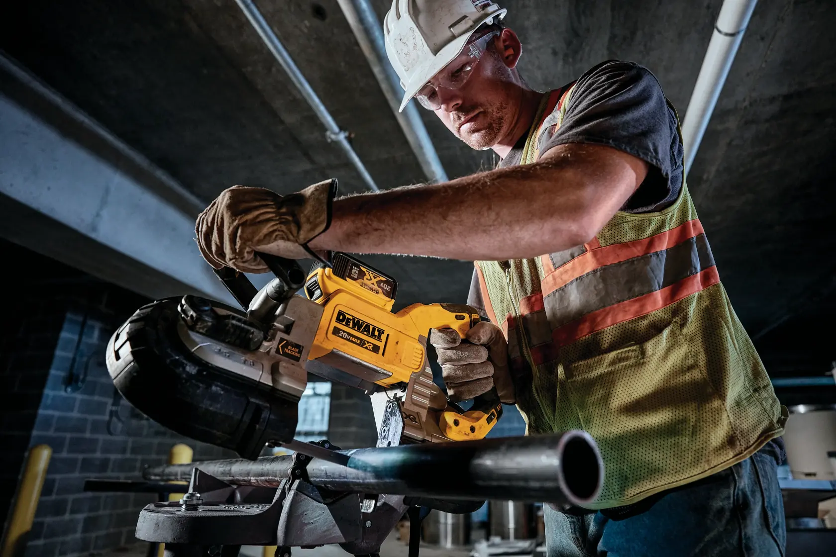 Um trabalhador da construção civil usando luvas, colete de segurança e capacete utiliza uma serra de fita sem fio DEWALT para cortar um tubo de metal em um ambiente industrial.