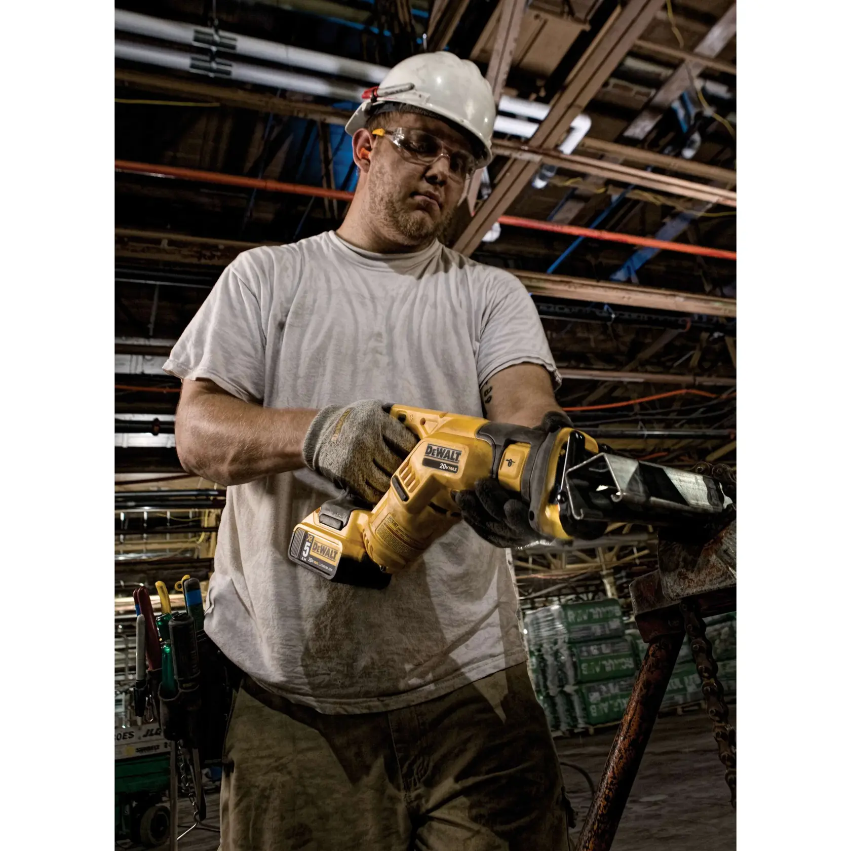 Uma pessoa usando luvas de trabalho e capacete está operando uma serra recíproca DEWALT DCS387B em um ambiente industrial. Ao fundo, há materiais de construção e tubos aparentes no teto.
