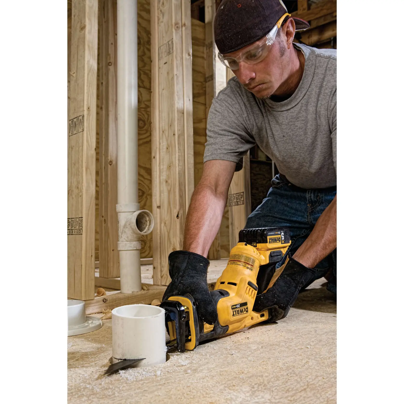 Uma pessoa usando luvas e boné está utilizando uma serra recíproca sem fio DEWALT para cortar um tubo de PVC em um canteiro de obras com estrutura de madeira e encanamento exposto.