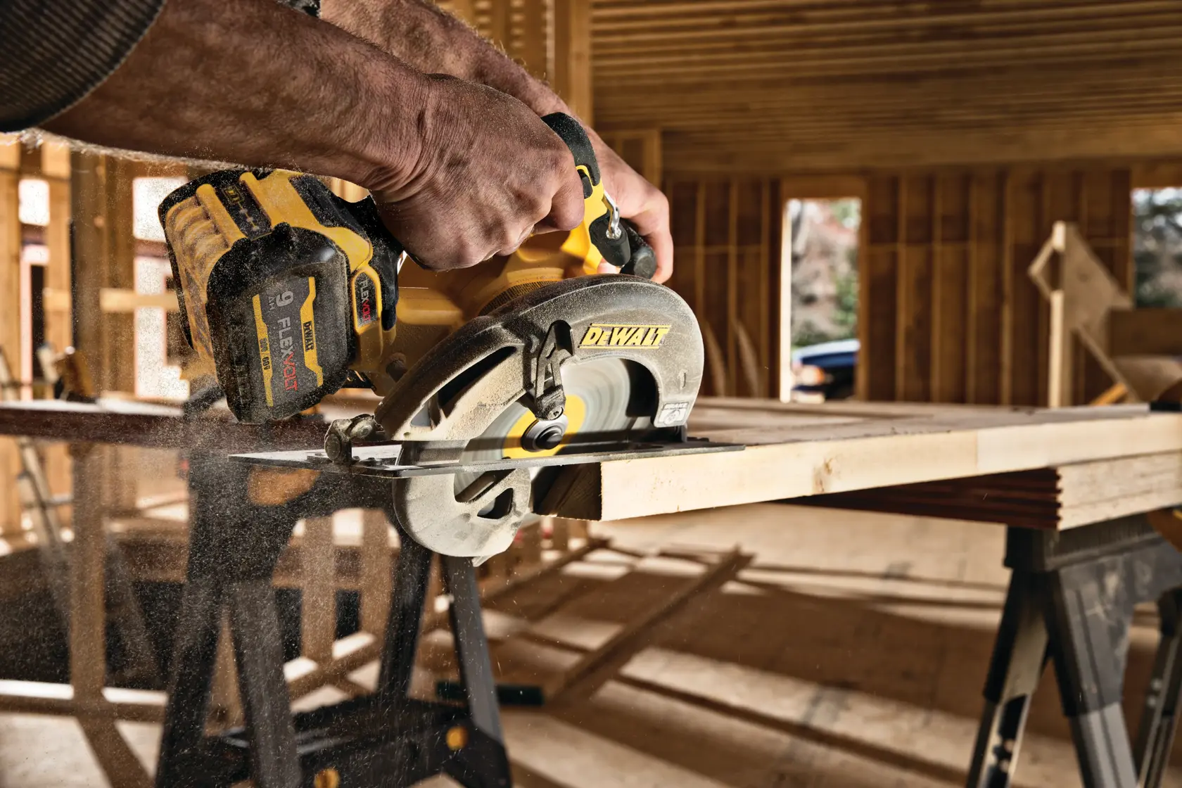 Una sierra circular inalámbrica DEWALT está siendo utilizada para cortar una tabla de madera dentro de un sitio de construcción. La sierra está en funcionamiento, se puede ver polvo de madera, y el fondo muestra una estructura de madera sin terminar.