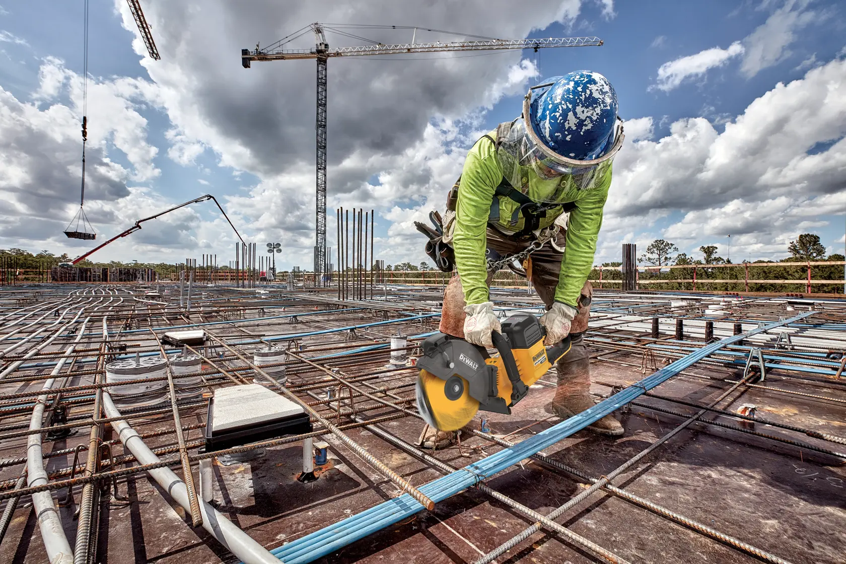 Um trabalhador da construção civil usando equipamentos de segurança está usando uma serra sem fio DEWALT para cortar um tubo ou conduíte em um canteiro de obras cheio de barras de aço e equipamentos de construção. Guindastes e um céu nublado são visíveis ao fundo.