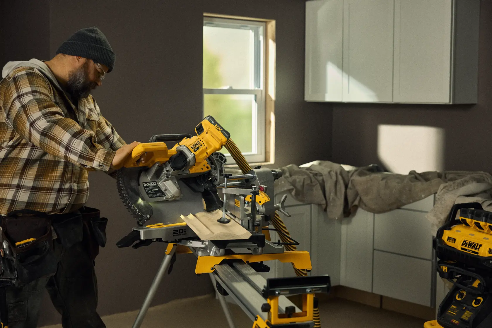 Uma pessoa opera uma serra de esquadria sem fio DEWALT (modelo DCS785B) para cortar tábuas de madeira em uma oficina ou sala renovada. O fundo apresenta armários, uma janela e outras ferramentas e equipamentos DEWALT.