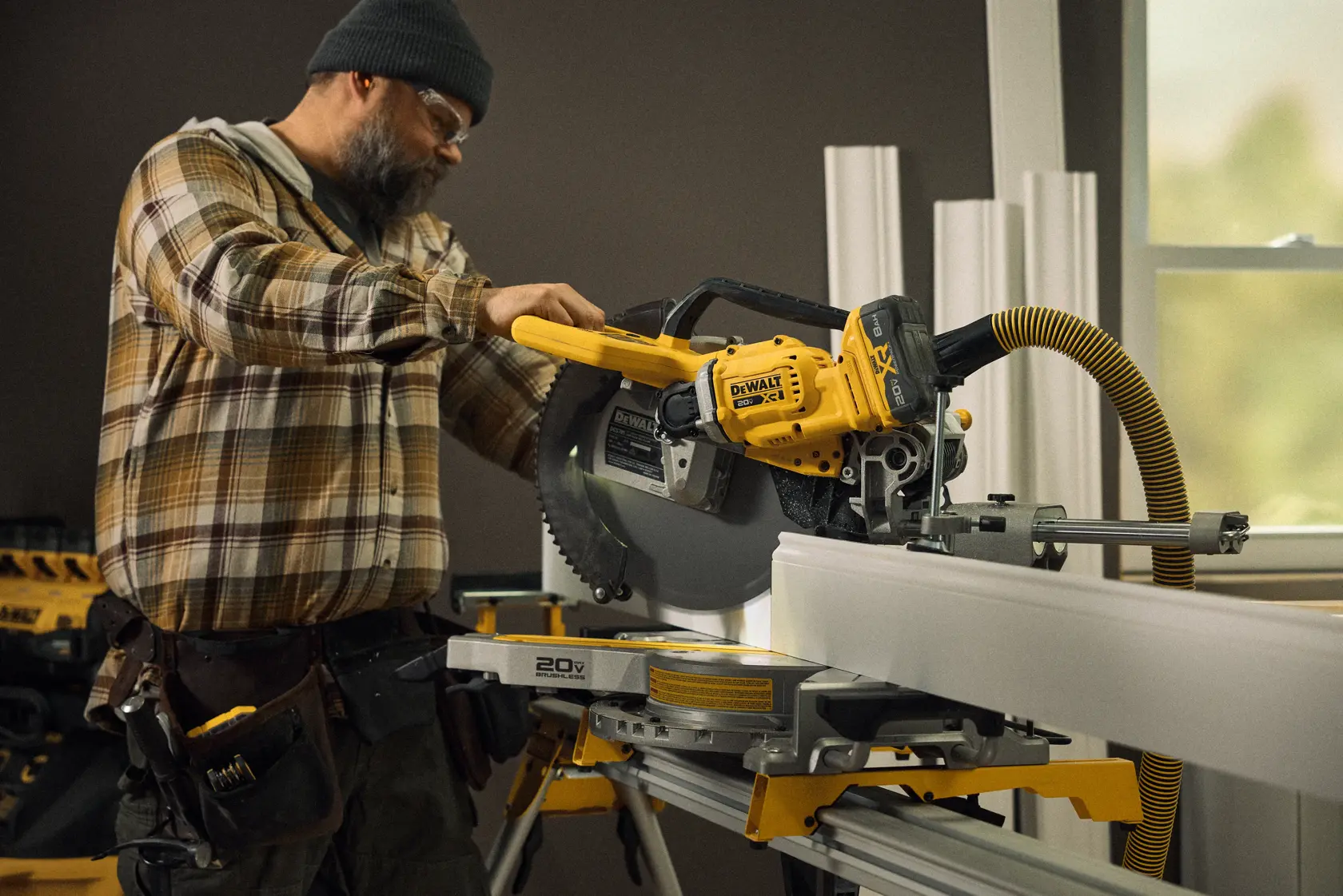 Uma pessoa usando uma serra de esquadria sem fio DEWALT DCS785B para cortar uma moldura em ambiente interno, com ferramentas de carpintaria visíveis no cinto de ferramentas e uma mangueira de extração de pó conectada à serra.