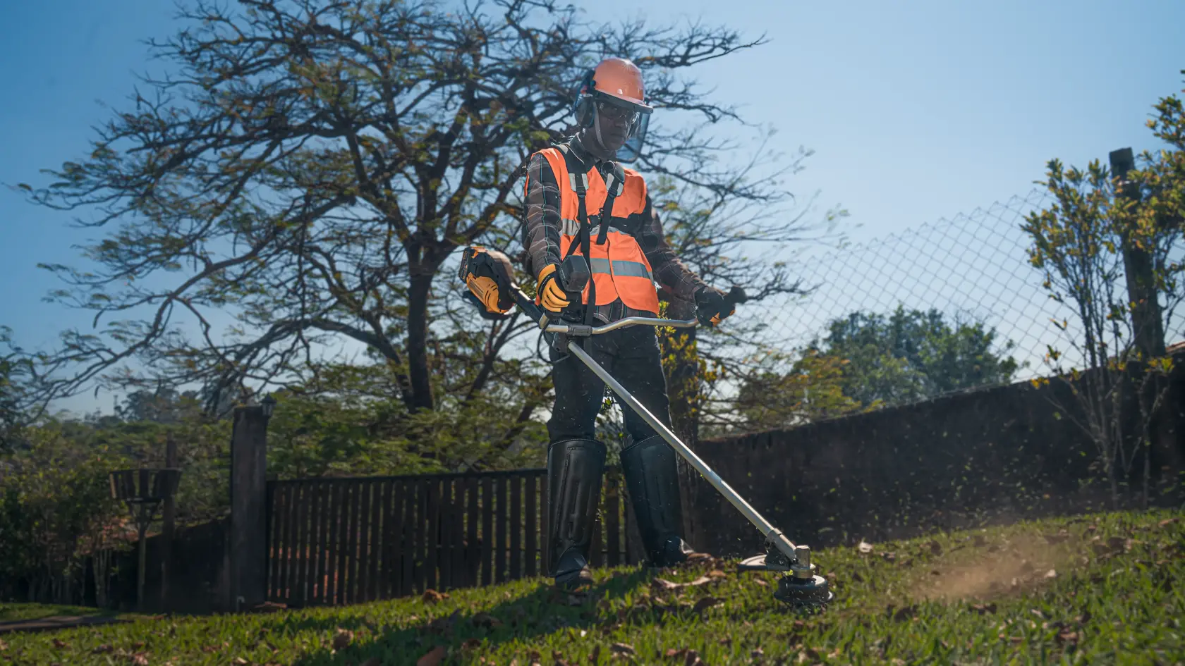 Uma pessoa usando equipamento de proteção, incluindo colete de segurança laranja, capacete, luvas e protetores de pernas, opera um aparador de grama para cortar a grama ao ar livre perto de uma cerca e árvores.