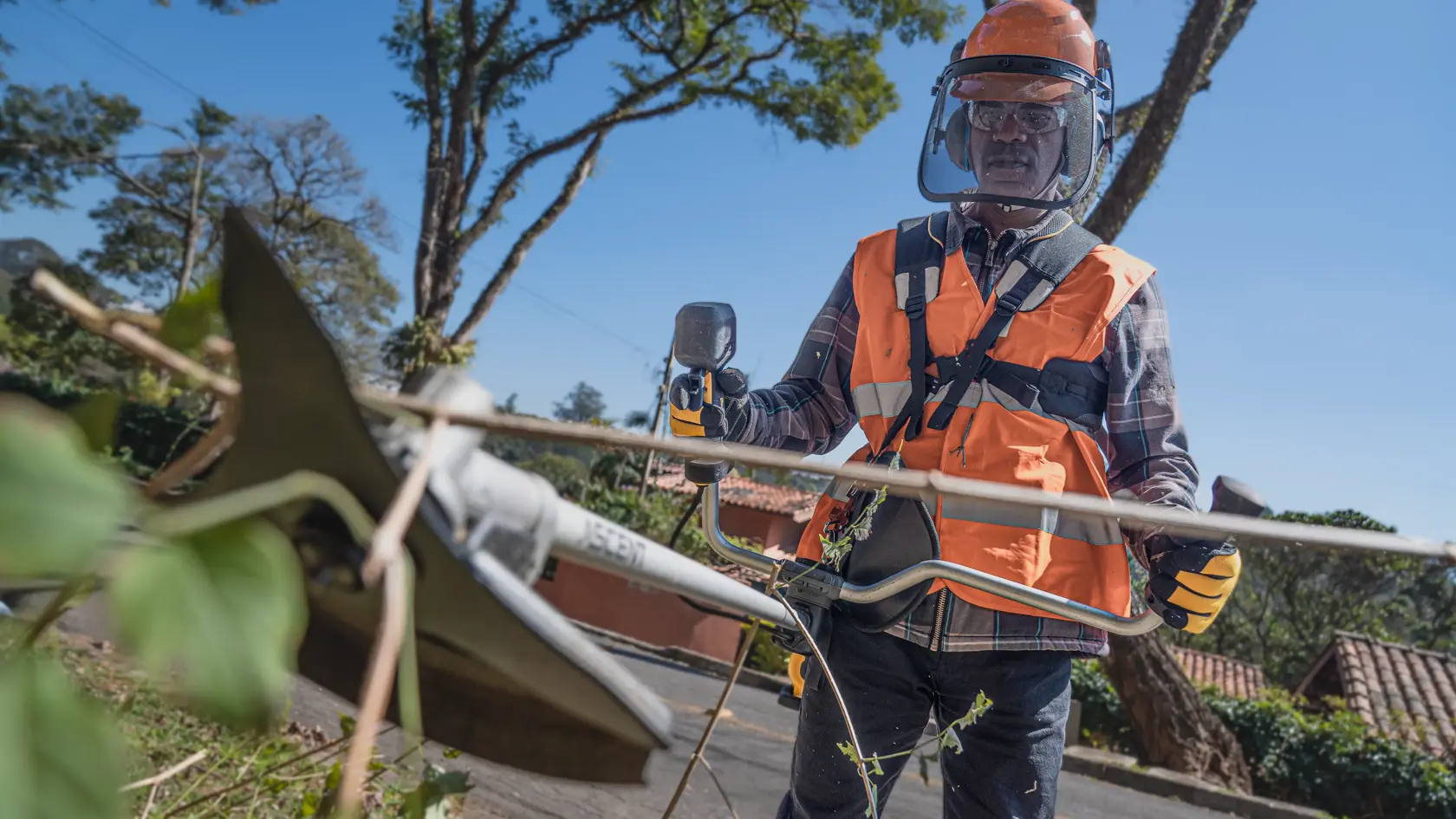 Uma pessoa usando equipamento de proteção, incluindo colete laranja, luvas e capacete com viseira, utiliza um cortador de grama DEWALT ao ar livre para cortar vegetação.