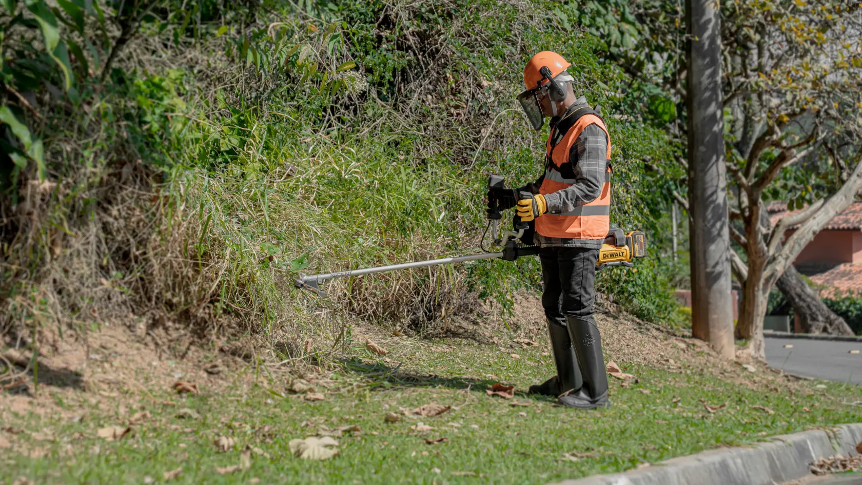 Uma pessoa usando equipamento de proteção opera um cortador de grama sem fio DEWALT para aparar grama e vegetação crescida perto de uma estrada.