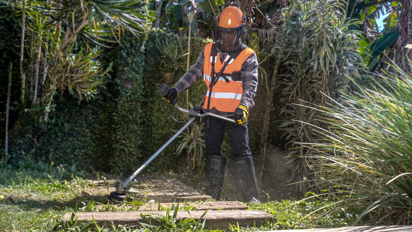Uma pessoa usando colete de segurança laranja, capacete de proteção, luvas e botas utiliza um cortador de grama DEWALT para aparar a grama em uma área de jardim cercada por folhagem verde.