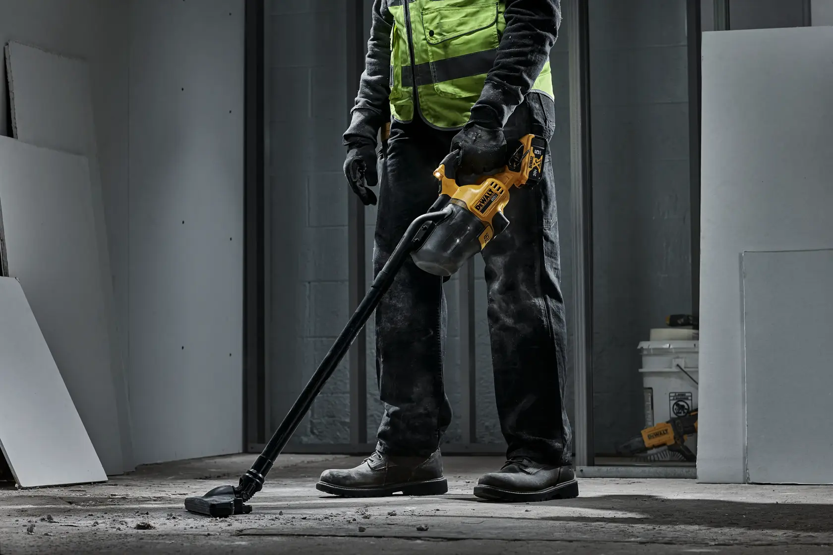 Uma pessoa vestindo colete de segurança de alta visibilidade e luvas está usando um aspirador portátil sem fio amarelo e preto da DEWALT para limpar poeira e detritos do piso de concreto em um ambiente em construção.