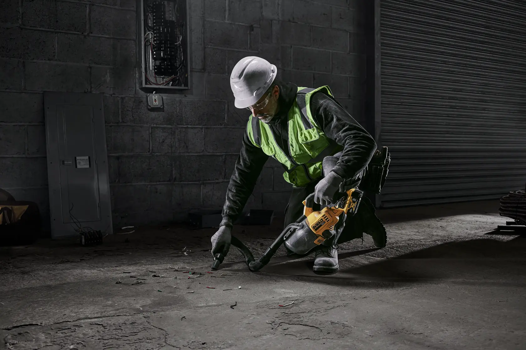 Um trabalhador usando colete de segurança e capacete está ajoelhado e usando um aspirador sem fio amarelo DEWALT para limpar detritos de um piso de concreto em um ambiente industrial.
