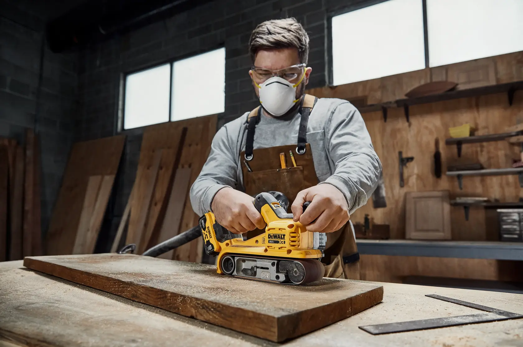 Uma pessoa está usando uma lixadeira de cinta amarela DEWALT 20V XR para lixar tábuas de madeira. O texto na parte inferior em francês diz: 'Placa de 3 pol x 21 pol oferece uma superfície de lixamento aumentada.'