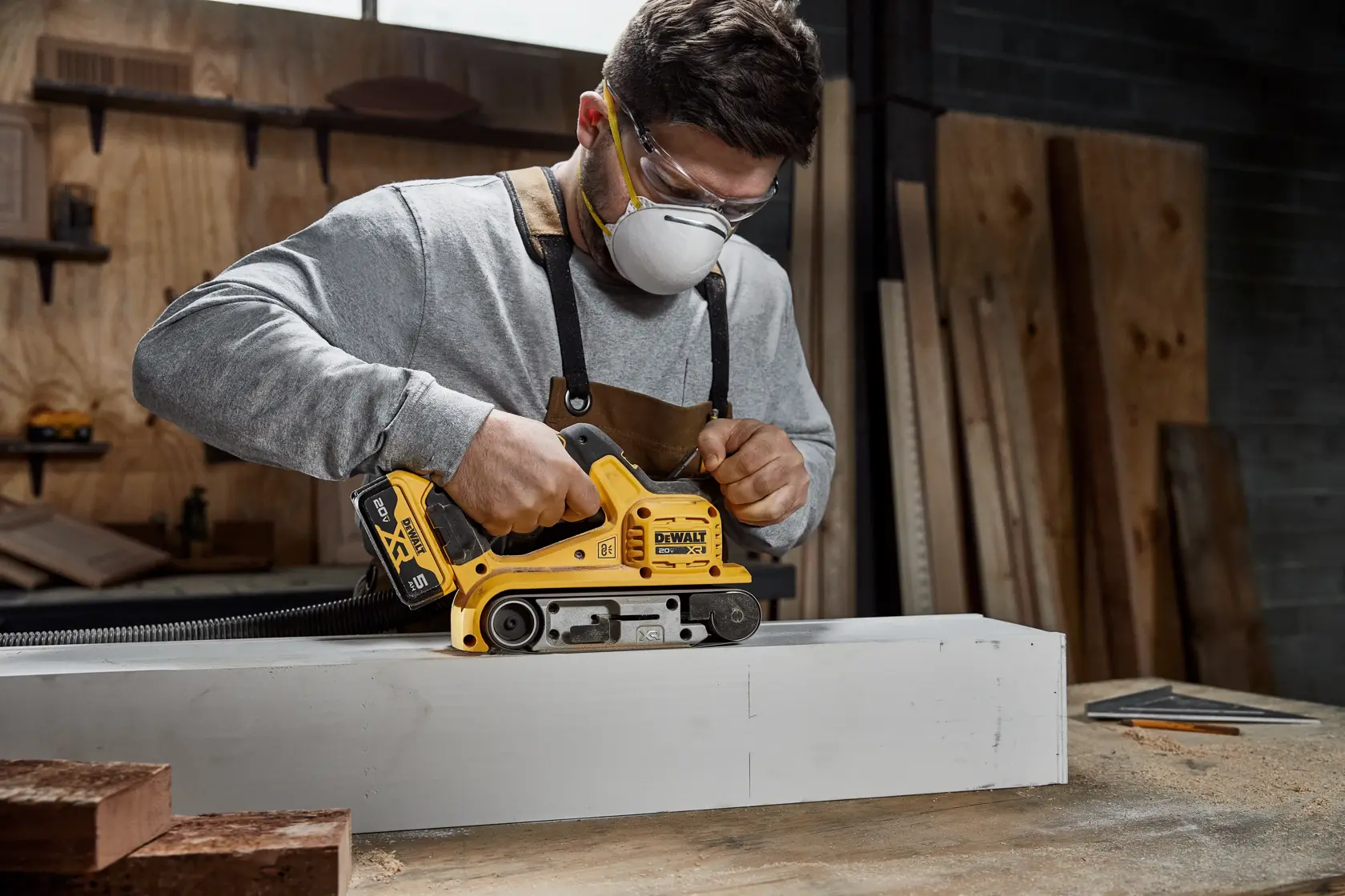 Uma pessoa usando uma lixadeira de cinta sem fio DEWALT 20V XR com uma mangueira de extração de poeira acoplada, lixando uma superfície de madeira. O texto destaca a minimização da poeira durante o lixamento e a compatibilidade com o sistema Airlock da DEWALT ou um extrator de poeira padrão de 35 mm. Bateria vendida separadamente.
