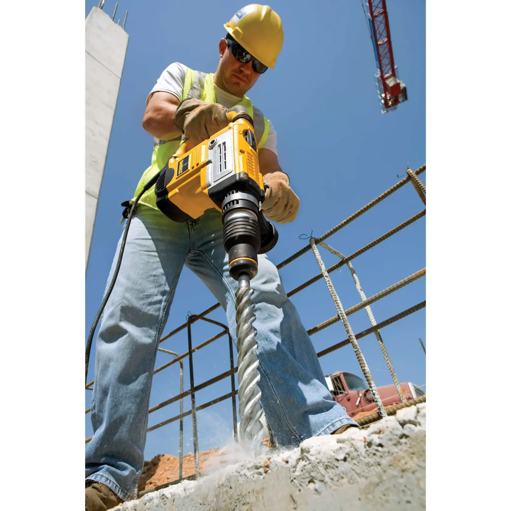 Um trabalhador da construção civil usa um martelete rotativo DEWALT com uma grande broca para perfurar concreto em um canteiro de obras ao ar livre. O trabalhador está usando capacete de segurança, luvas, colete refletivo e jeans. Barras de aço e um guindaste são visíveis ao fundo sob um céu azul claro.