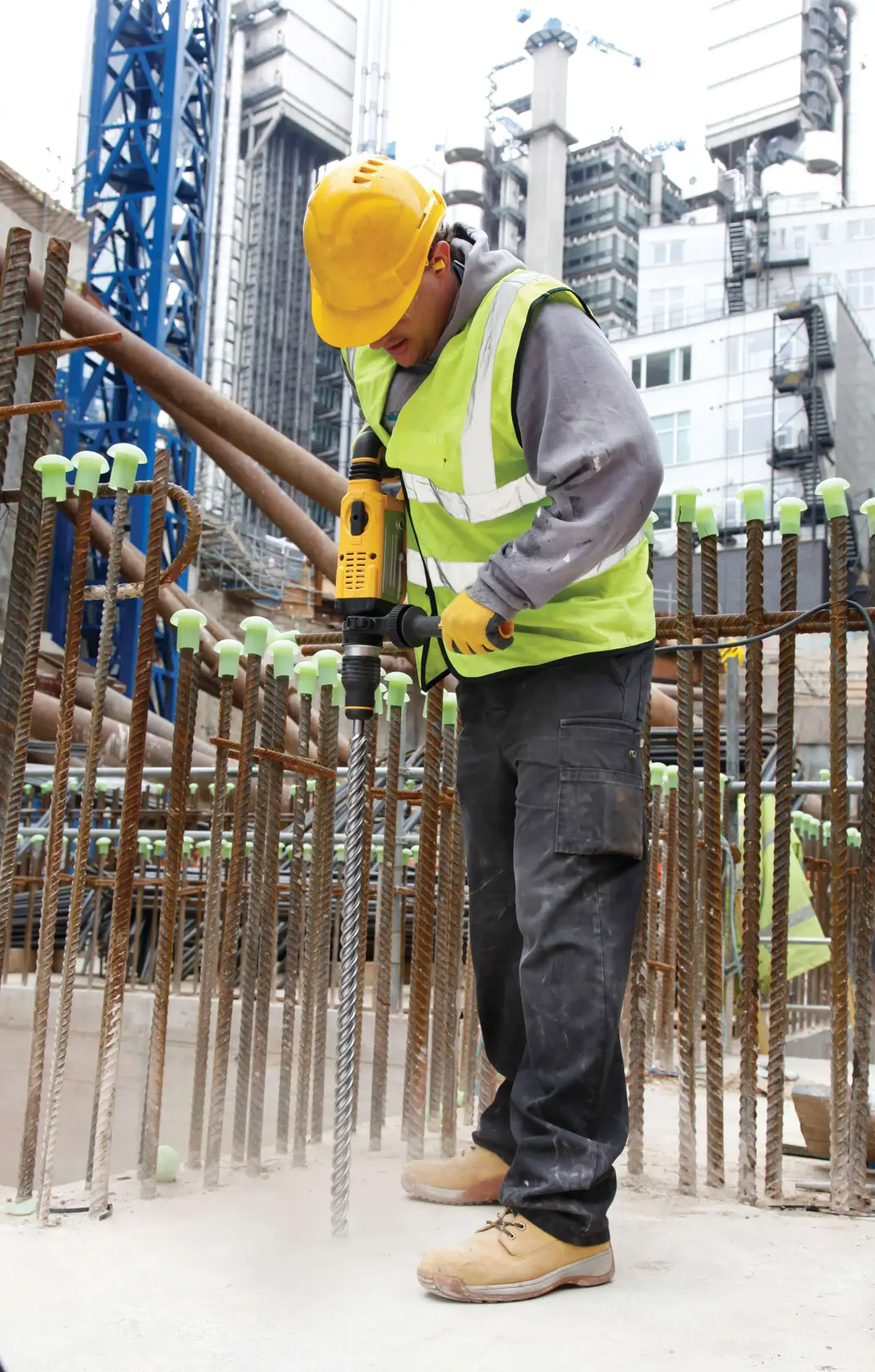 Um trabalhador da construção civil usando capacete amarelo, colete de alta visibilidade, luvas e botas de trabalho utiliza um martelete rotativo DEWALT para trabalhar no concreto em um canteiro de obras. Ele está cercado por vergalhões de aço e edifícios urbanos são visíveis ao fundo.