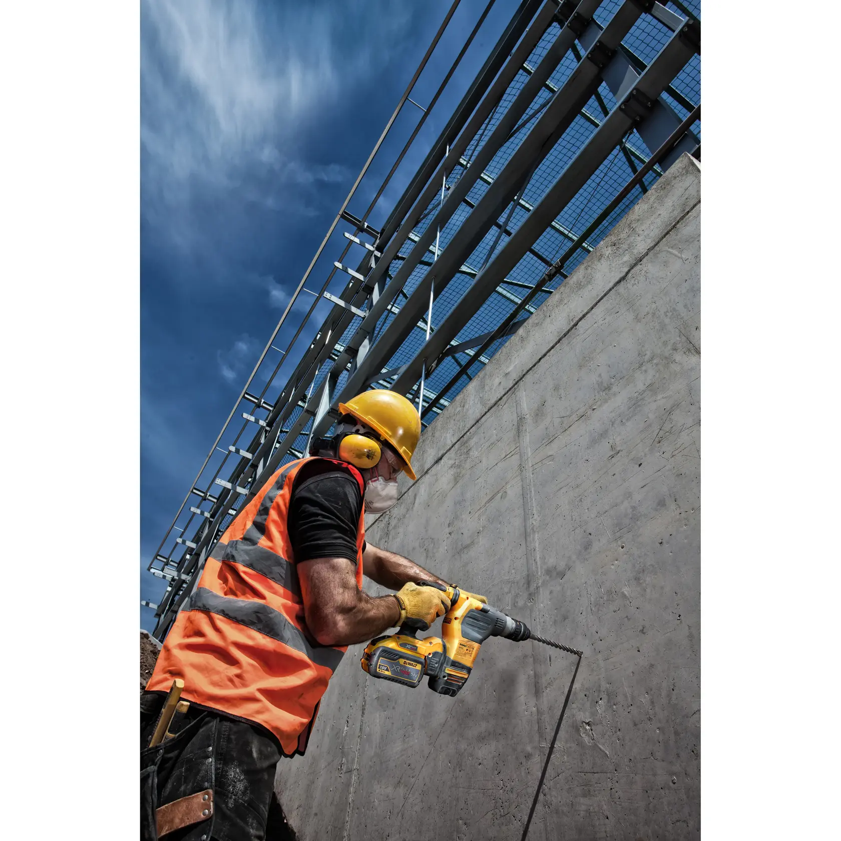Trabalhador com equipamento de segurança usa furadeira Dewalt em parede de concreto sob céu azul.