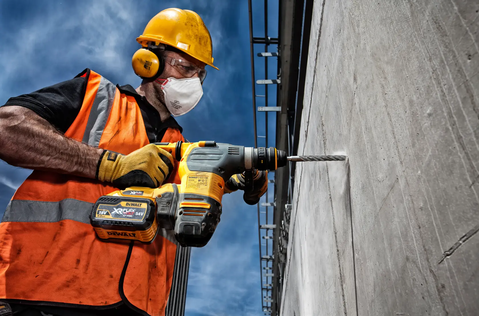Trabalhador usa martelo DEWALT para furar parede de concreto ao ar livre, com equipamento de segurança.