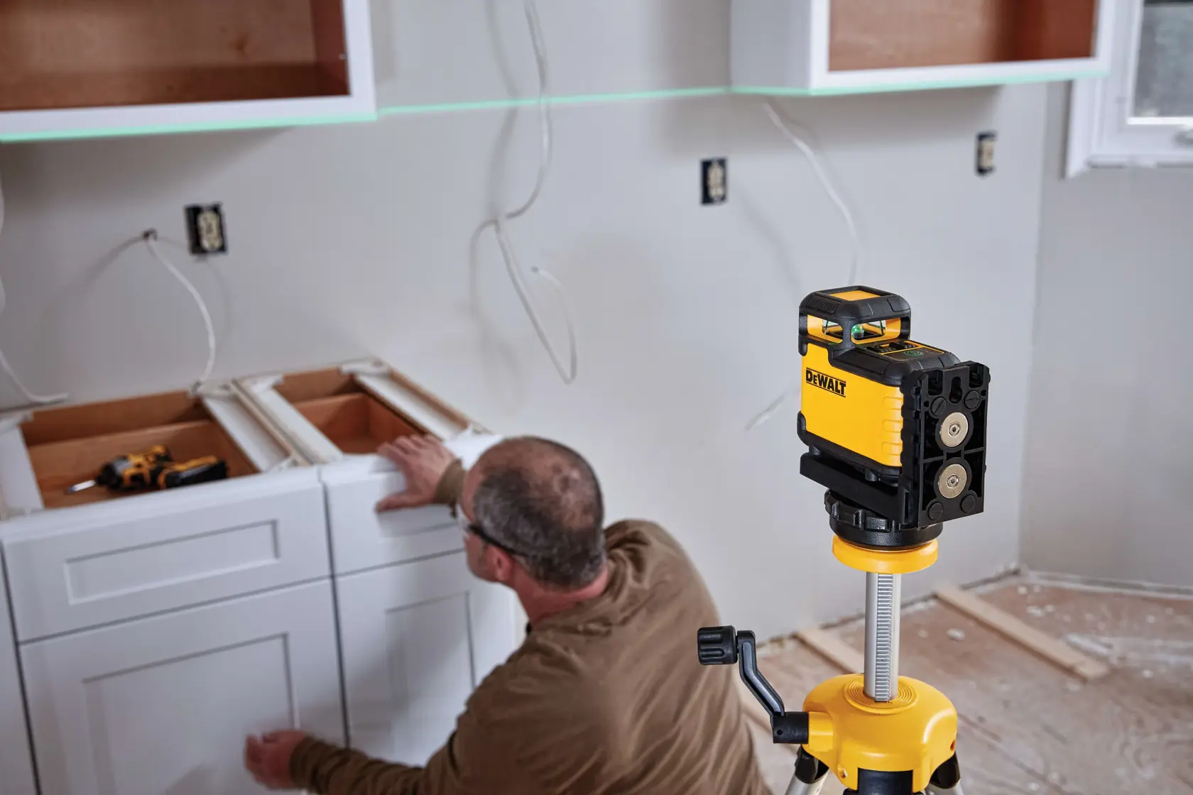 Um nível a laser verde DEWALT está montado em um tripé em uma sala em reforma, projetando uma linha verde na parede. Uma pessoa com o rosto desfocado aparece ao fundo, perto de um armário e área de trabalho.