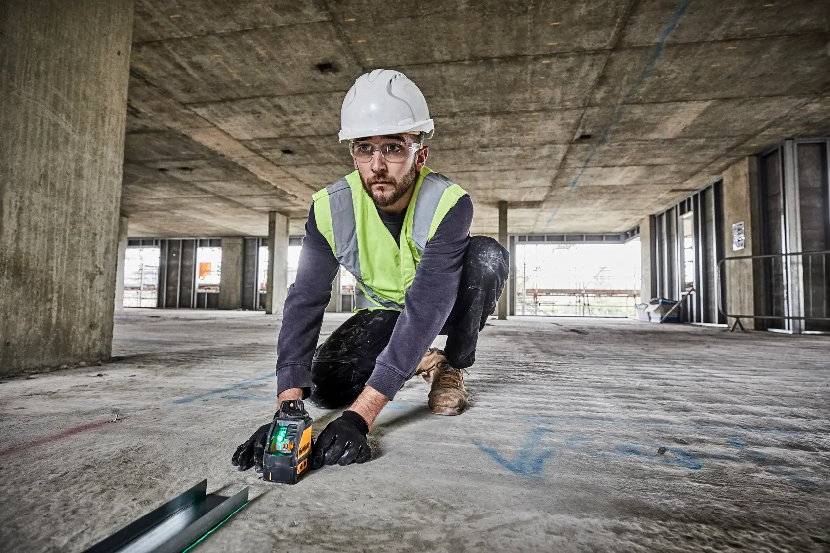 Foto de trabalhador usando DEWALT DW088CG laser em obra, ajoelhado no chão de concreto.