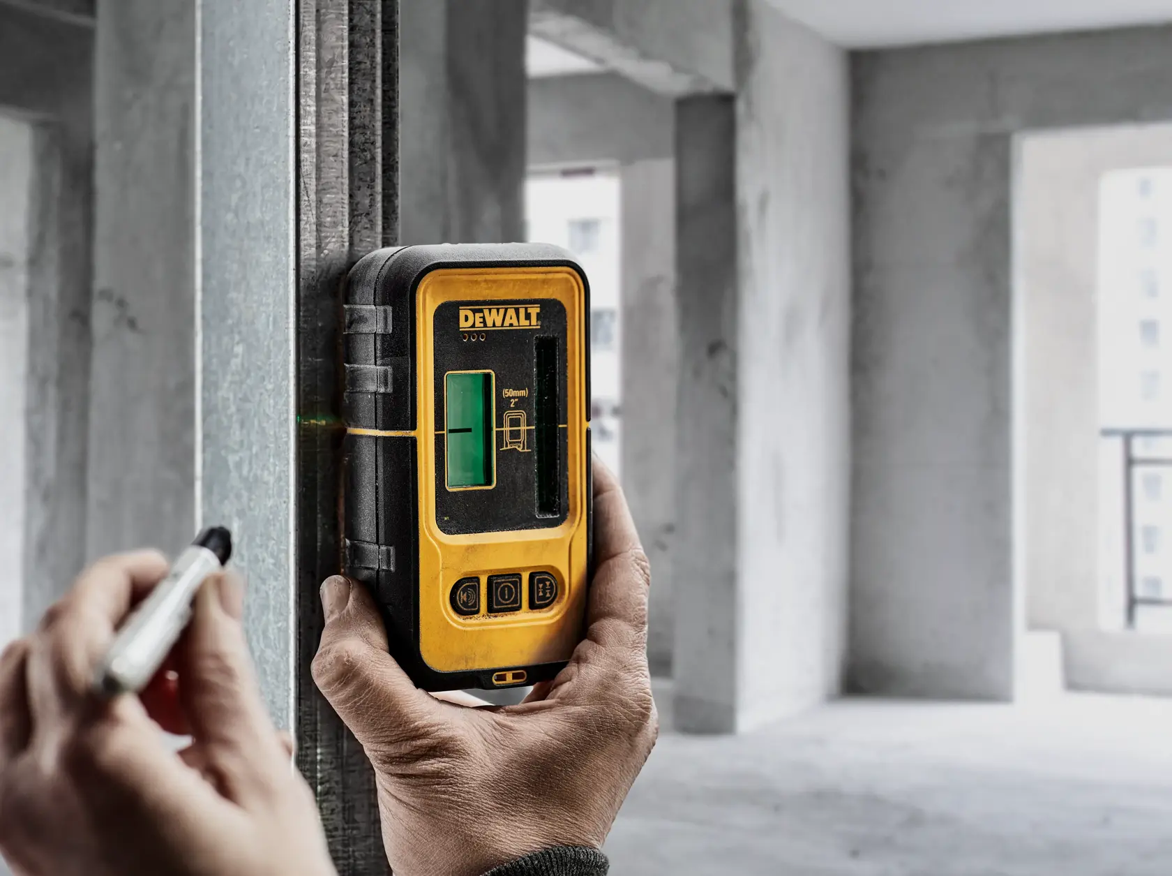 Uma pessoa usa um detector de laser verde DEWALT DW0892G preso a uma viga de metal em um canteiro de obras, marcando uma linha com um marcador.