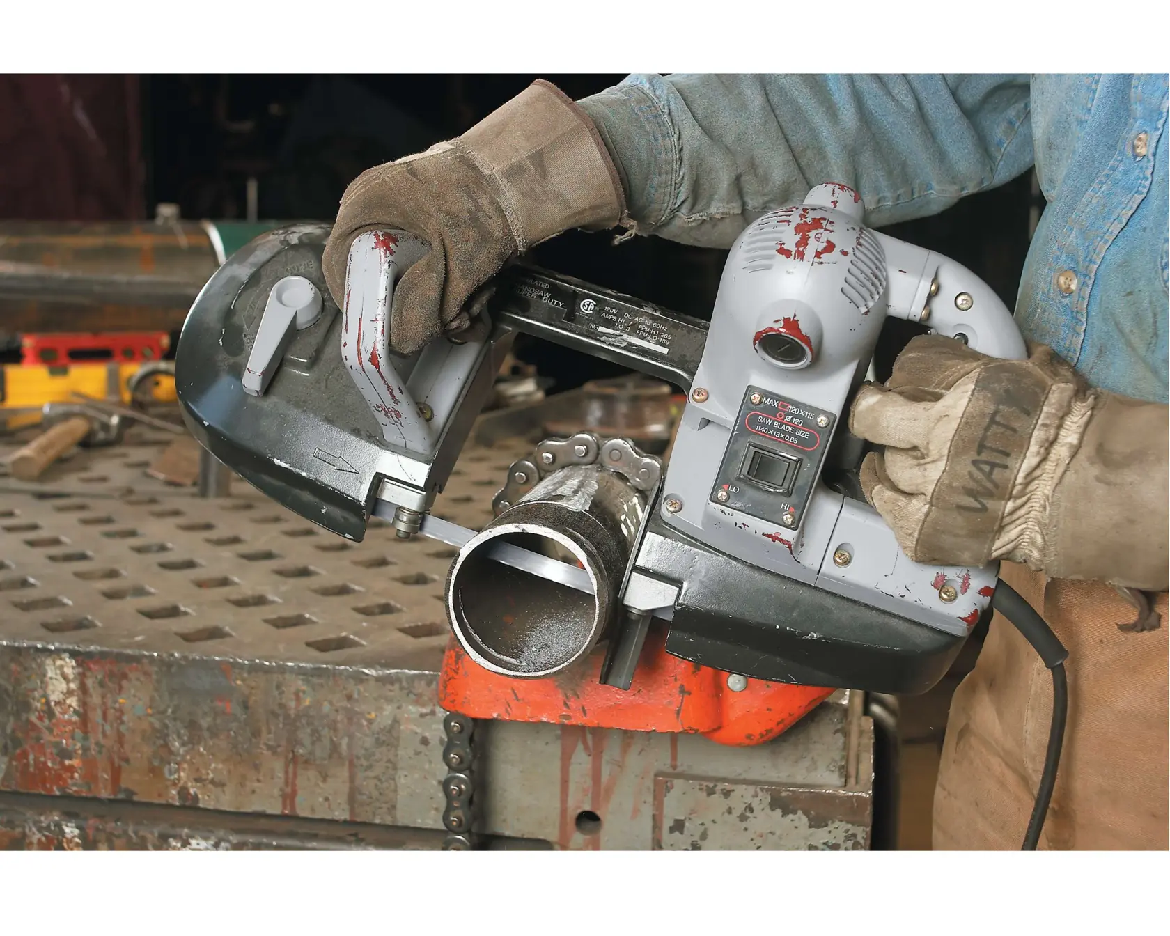 Uma pessoa usando luvas e camisa jeans utiliza uma serra de fita portátil DEWALT para cortar um tubo de metal em um ambiente de oficina industrial. A serra de fita está posicionada sobre o tubo, e ferramentas e equipamentos estão visíveis na superfície de trabalho.