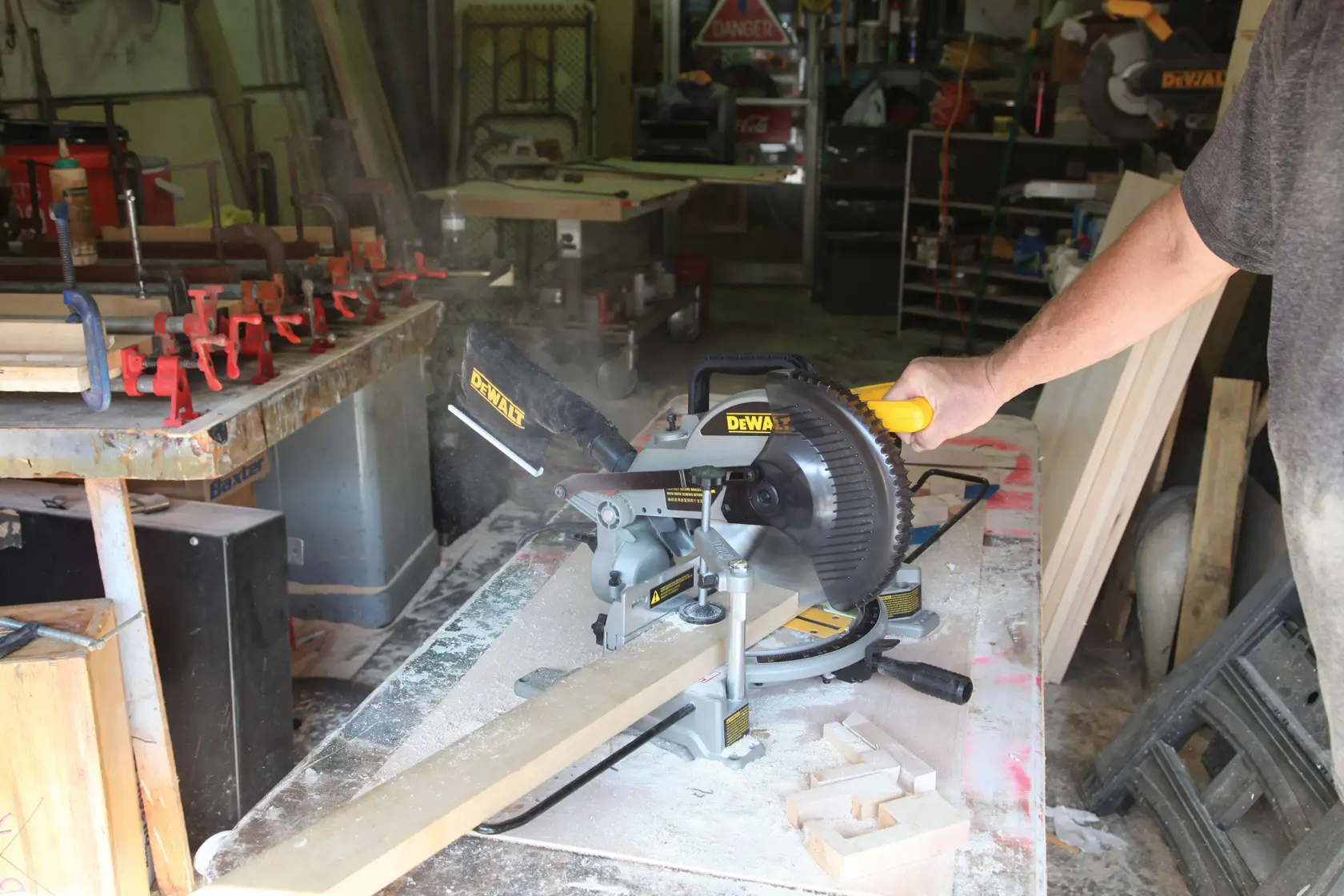 Uma pessoa opera uma serra de esquadria DEWALT (SKU: DW714) para cortar uma tábua de madeira em uma oficina. Poeira de madeira e grampos são visíveis na mesa de trabalho, com várias ferramentas e materiais ao fundo.