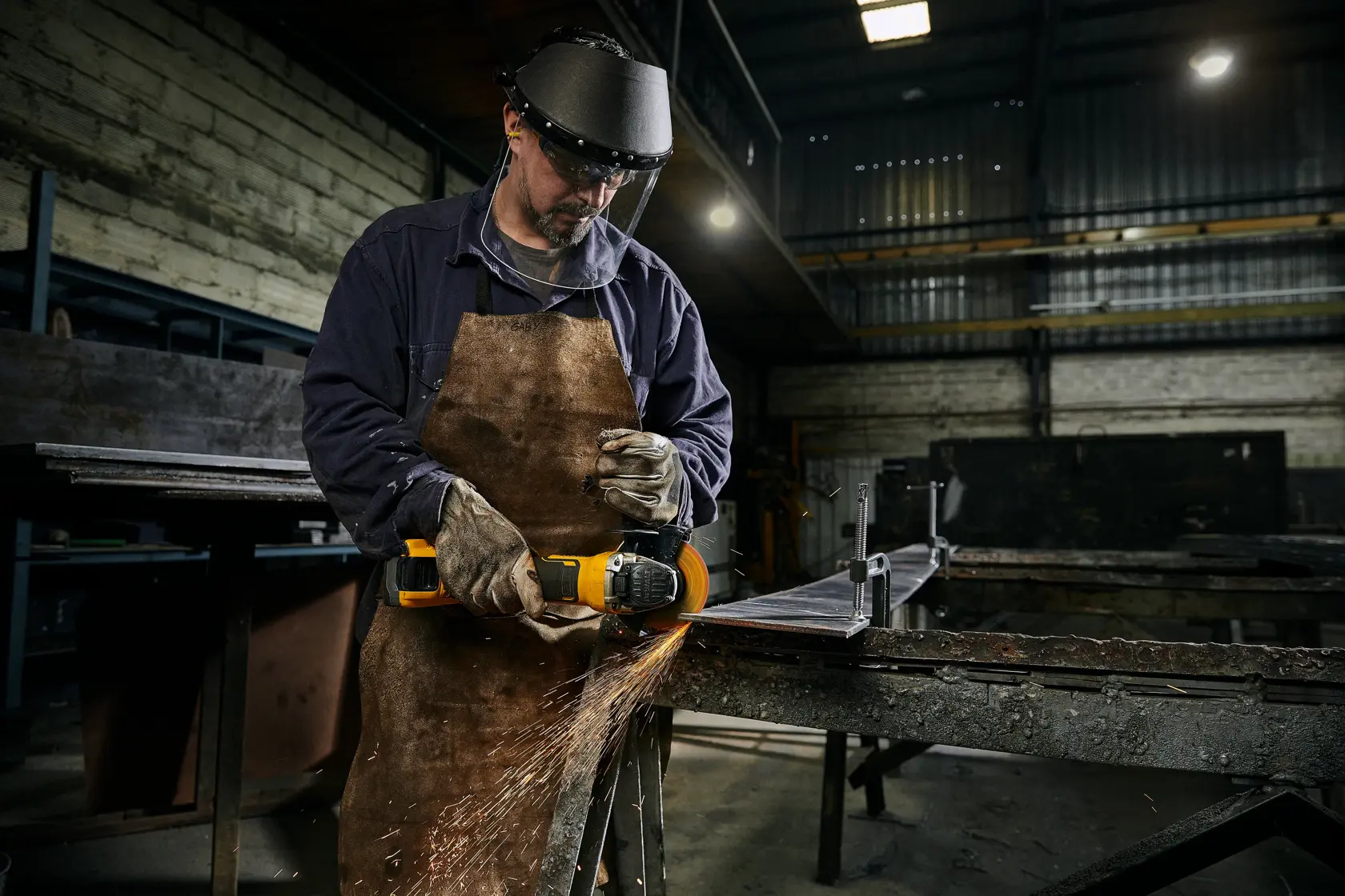 Trabalhador usa esmerilhadeira DEWALT, com equipamento de segurança e avental, gerando faíscas em oficina.
